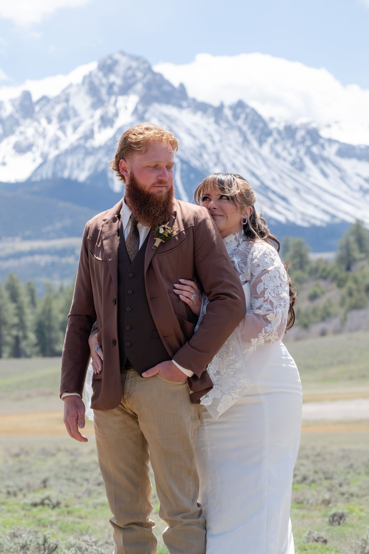 A bride stands behind her groom and embraces in front of snow-capped mountains, groom in brown suit with beard, bride in lace wedding dress.