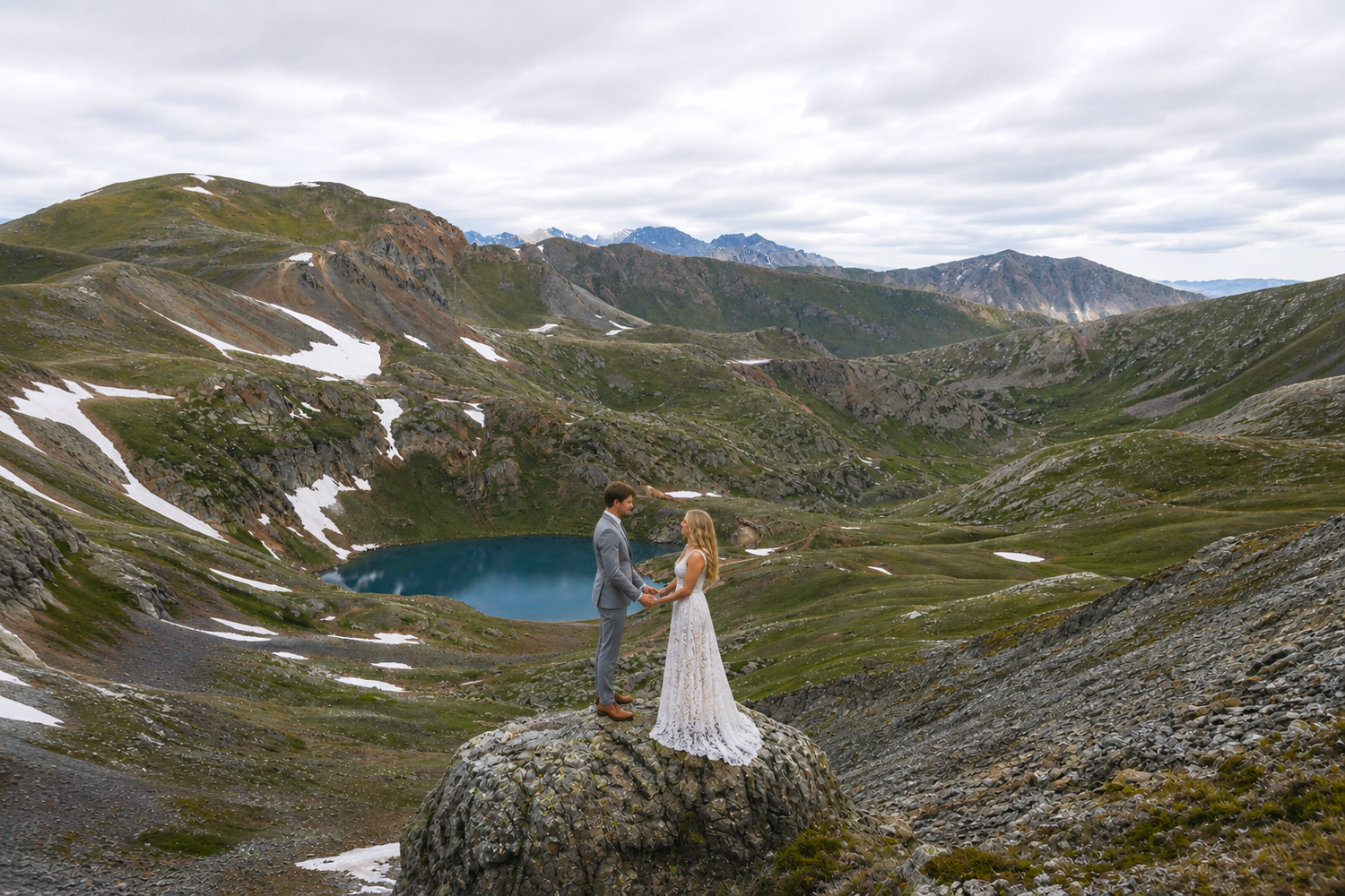 Couple holding hands on a rocky peak with a turquoise alpine lake and snow-dusted peaks of the San Juan mountains in the background.