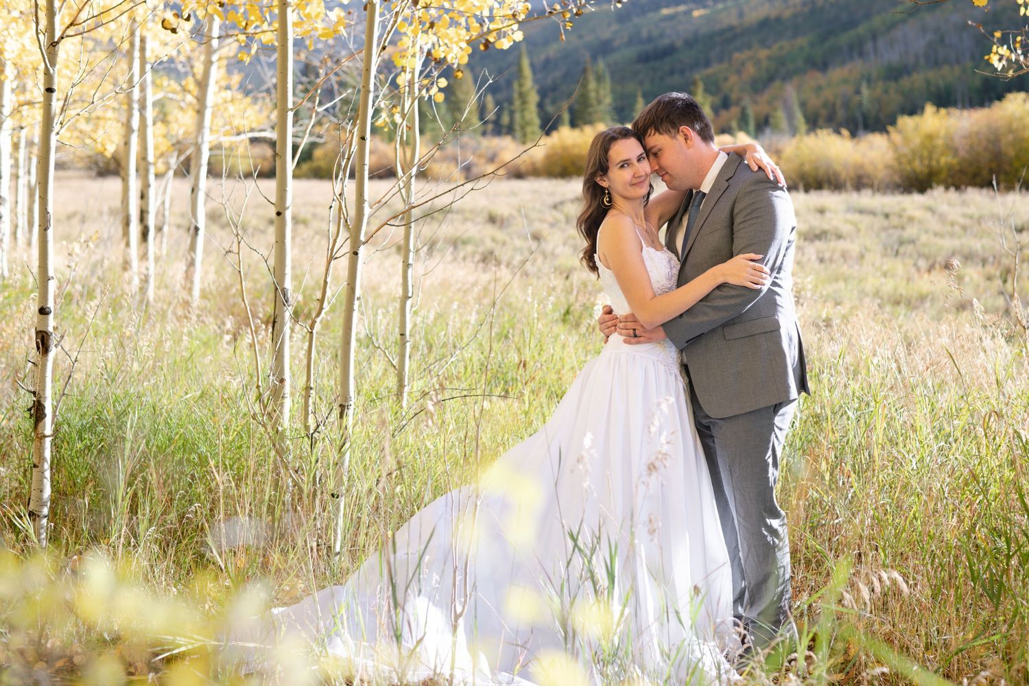 An eloping couple embracing in golden autumn meadow with aspen trees, bride in flowing white gown, groom in gray suit.