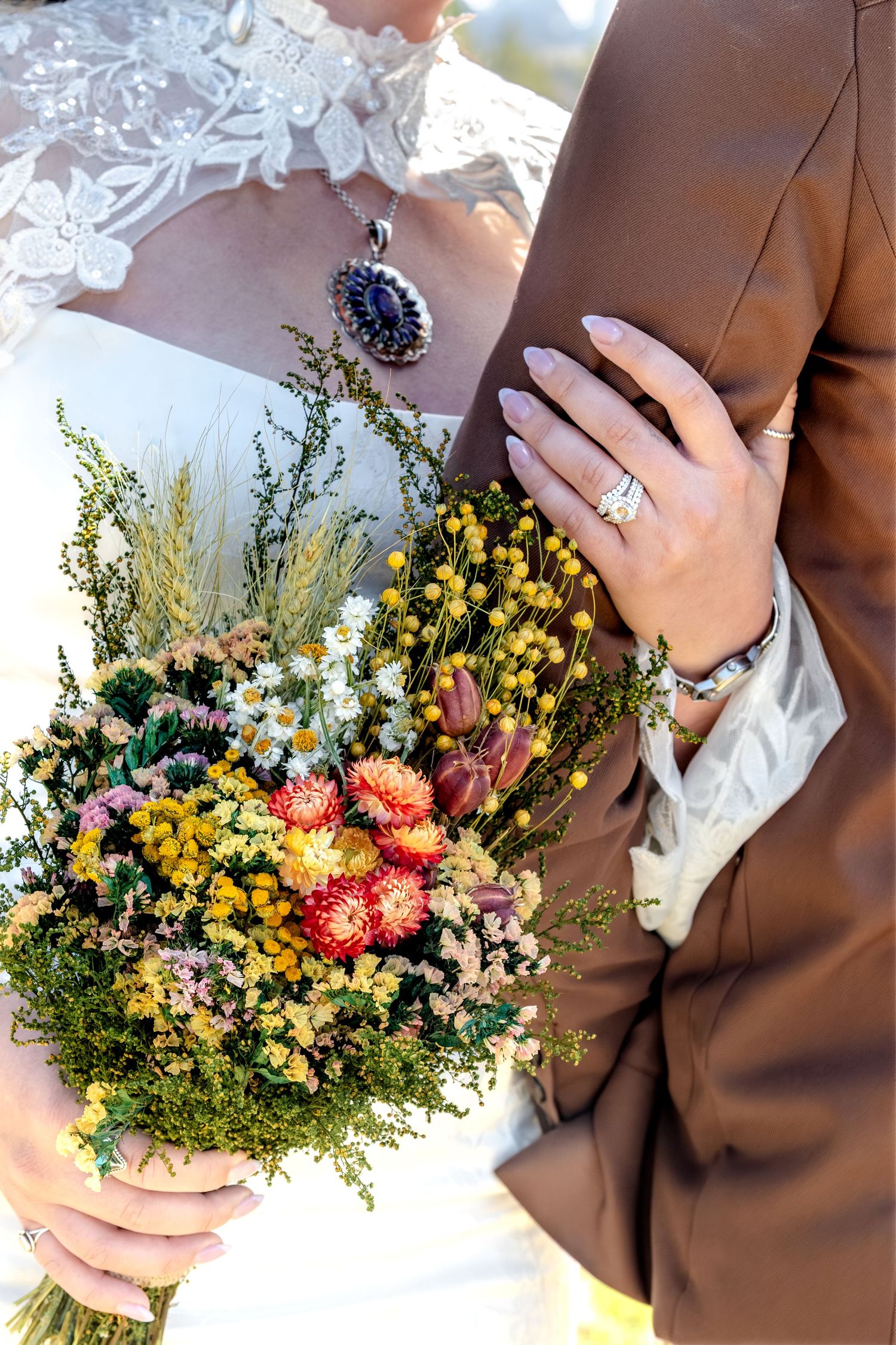 A detail image of a bride holding a colorful wildflower bouquet with groom in brown suit, wedding rings visible on her hand.