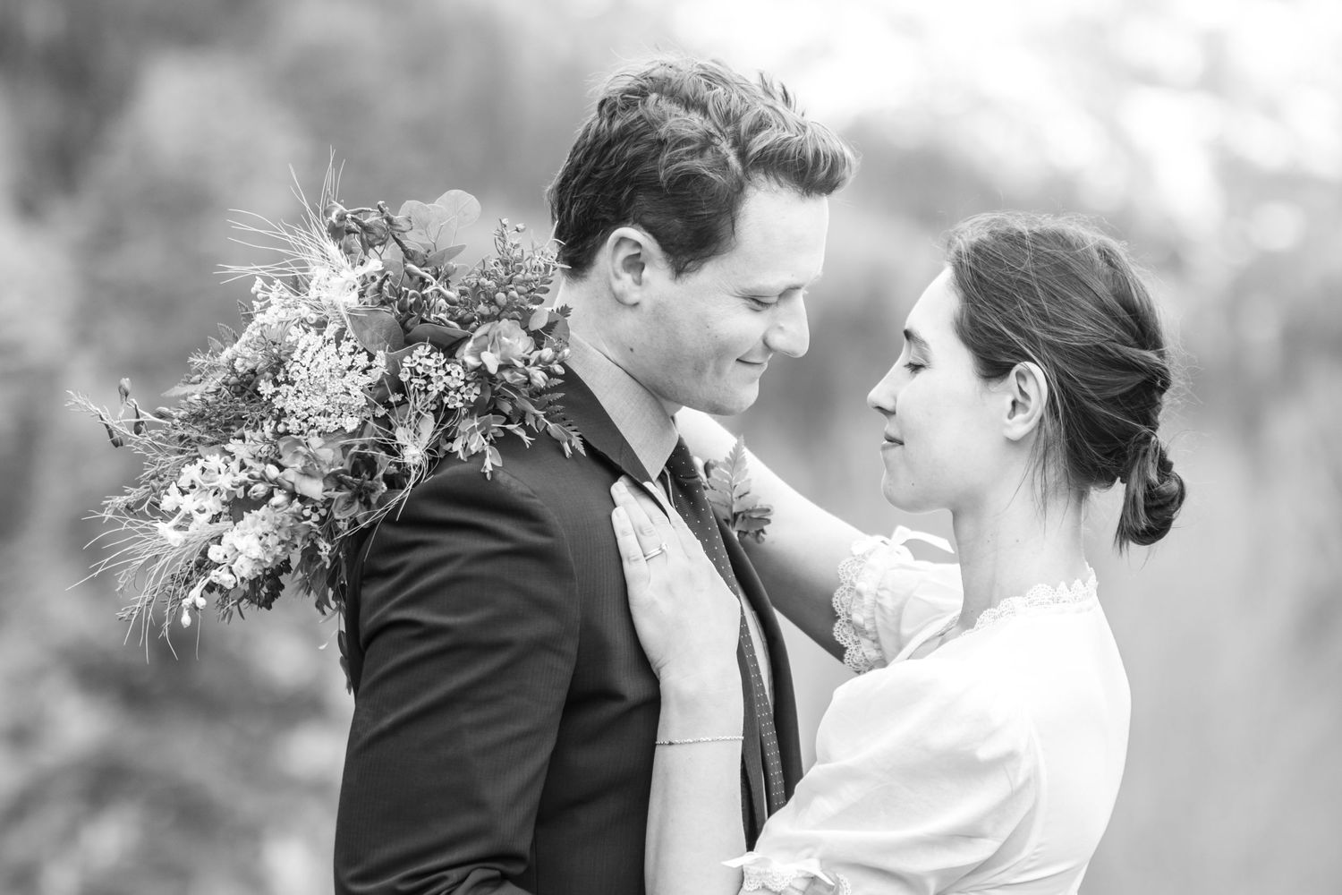 Black and white wedding photo of bride and groom in an intimate embrace. Bride is holding  the bouquet around her grooms shoulder outdoors.