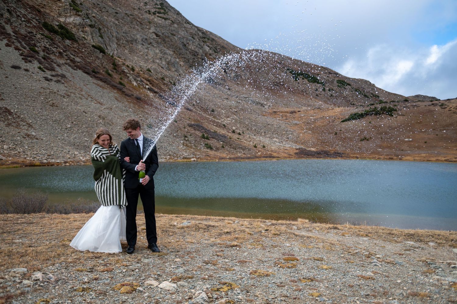 An eloping couple share a joyous moment of a champagne pop after saying their vows at a high alpine lake in the fall.