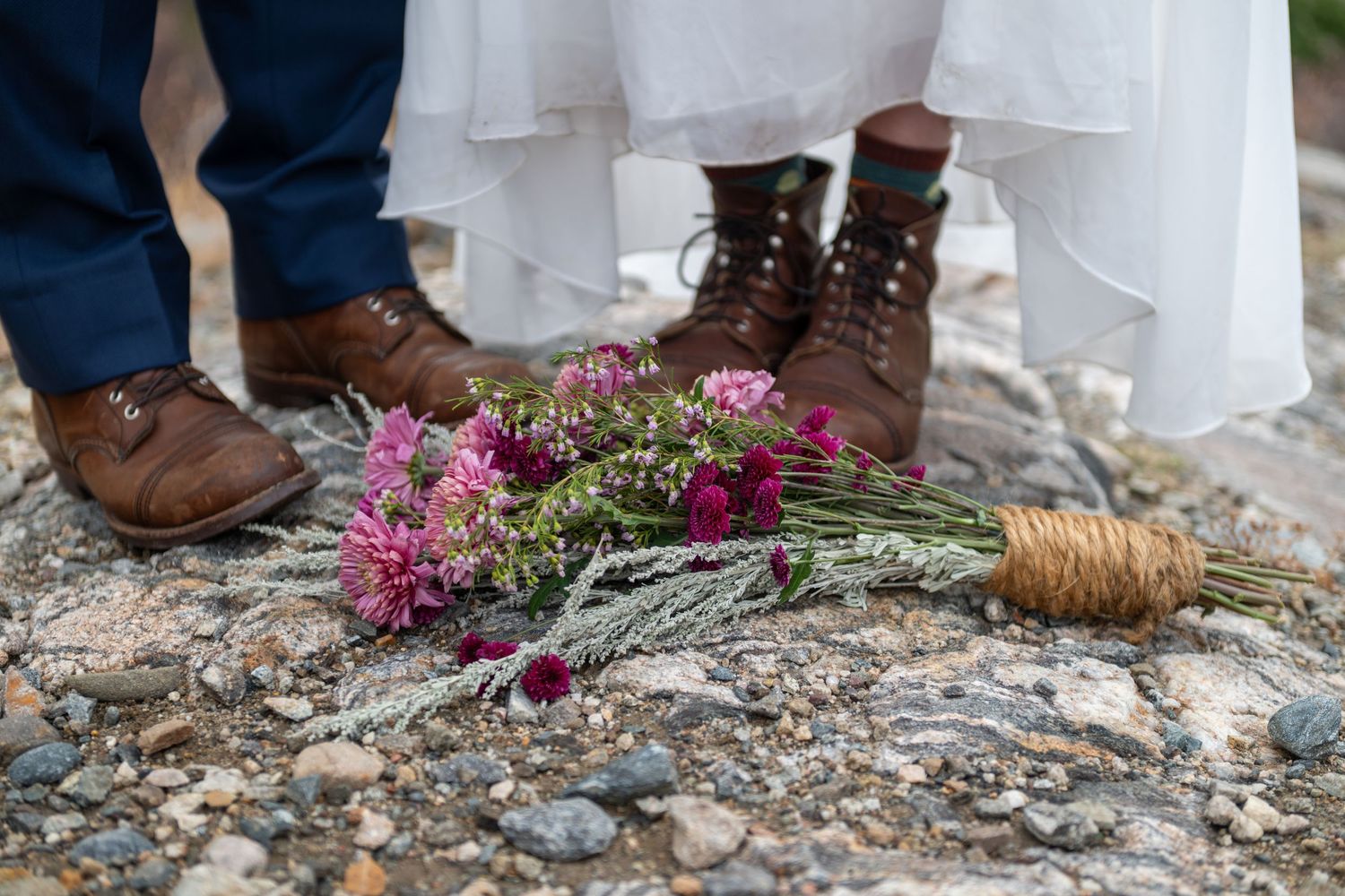 Bride and groom in brown boots standing over a pink wildflower bouquet and pinecone on rocky ground.