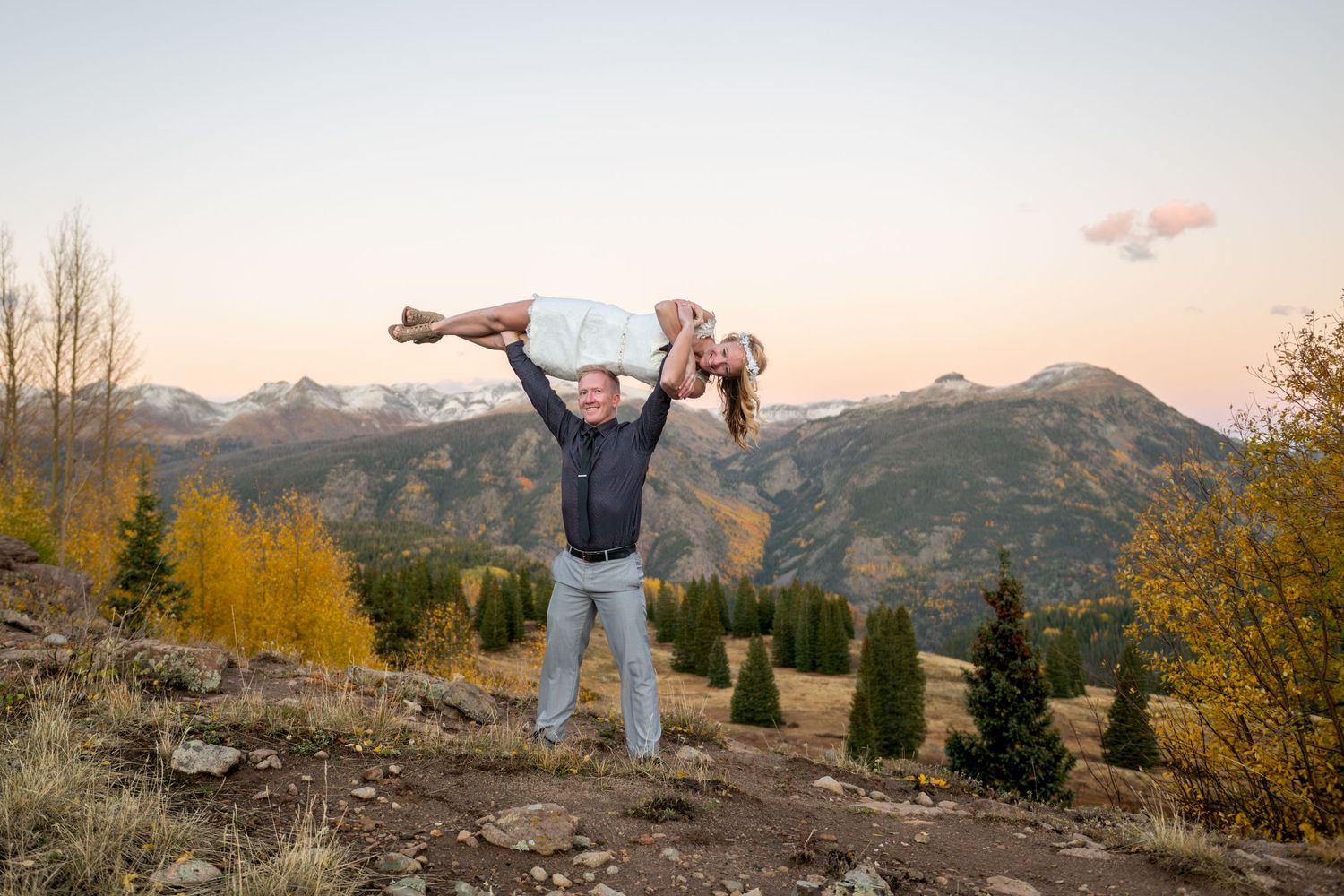A groom holds his bride overhead in a shoulder press with dramatic mountain peaks at sunset behind him. They are smiling and laughing.