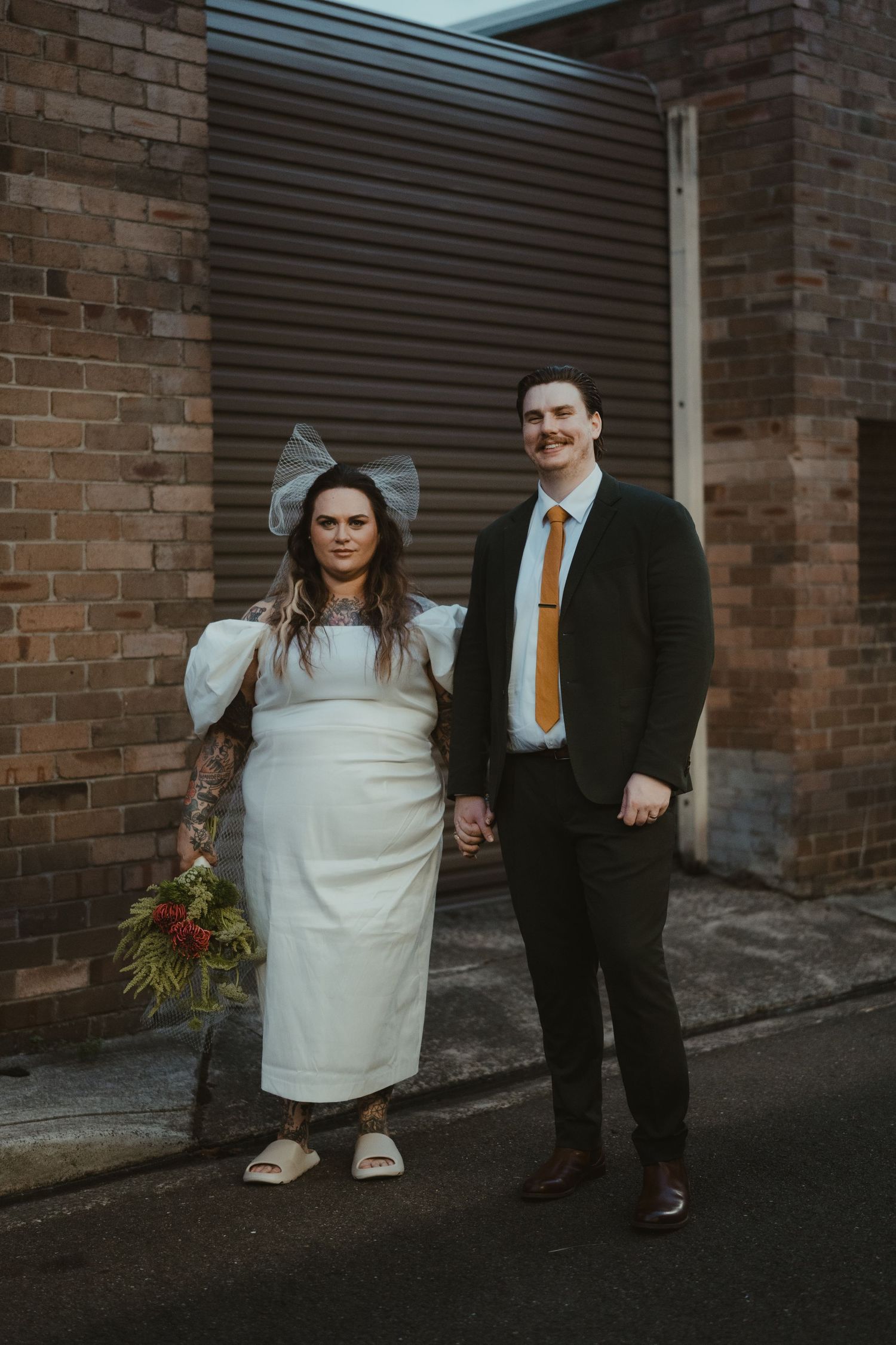 Wedding couple poses against industrial brick wall backdrop in urban setting.