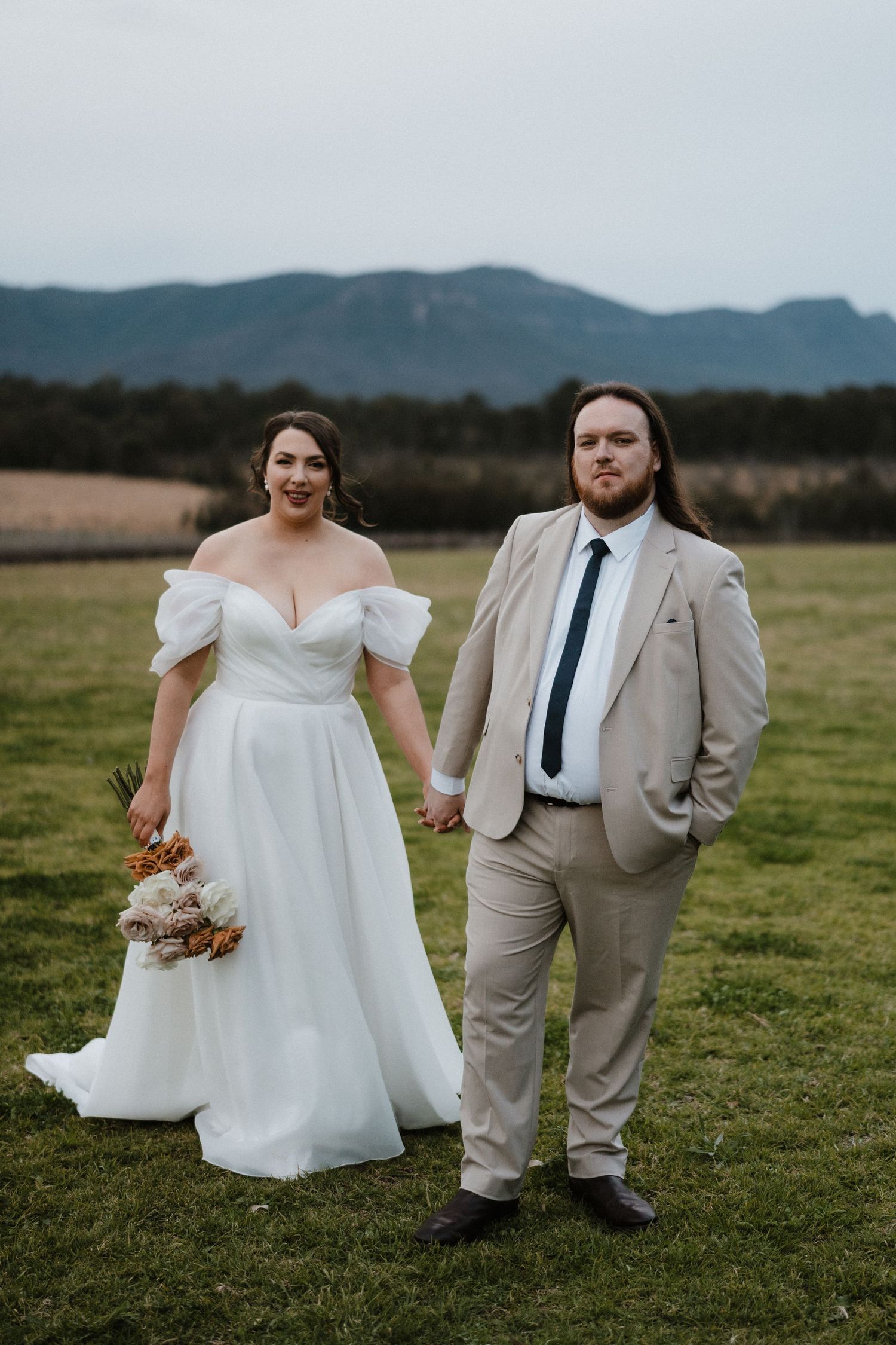Couple in wedding attire poses in field with mountains visible in background.