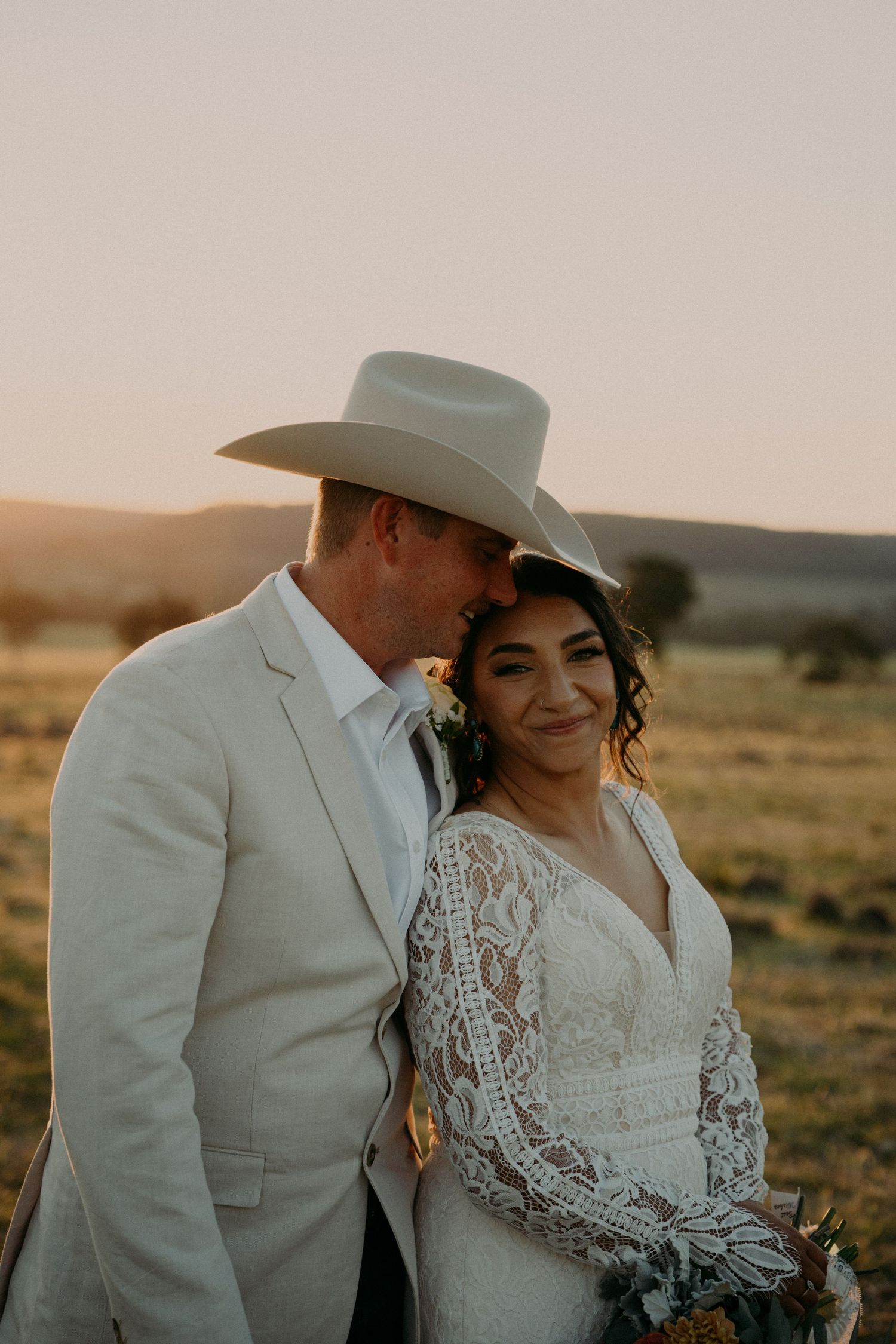 Bride in lace gown and groom in tan suit with cowboy hat share a romantic sunset moment in an open field.