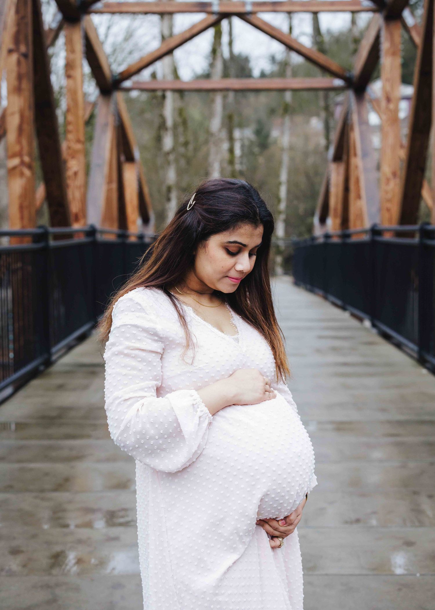 A pregnant woman holding her belly in a soft and sweet manner, at an outdoor park surrounded by trees near Seattle by Tim Neal Photography