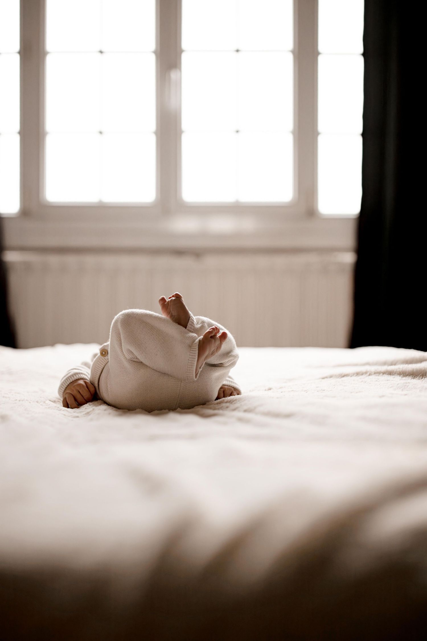 Small baby feet wrapped in white cloth on a soft bed near a bright window.