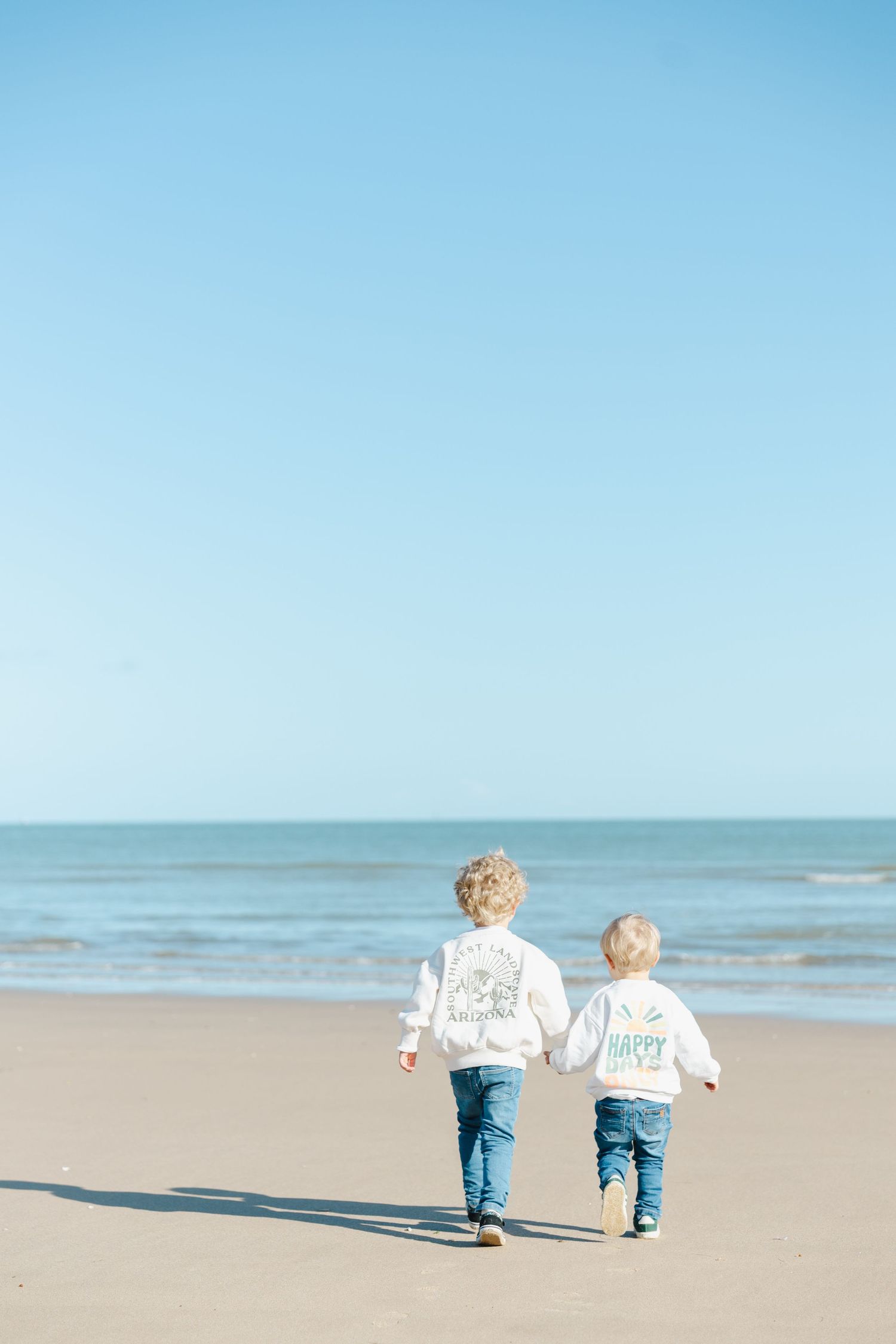 Two small children in matching white sweatshirts and jeans run together on a sunny beach by the ocean.