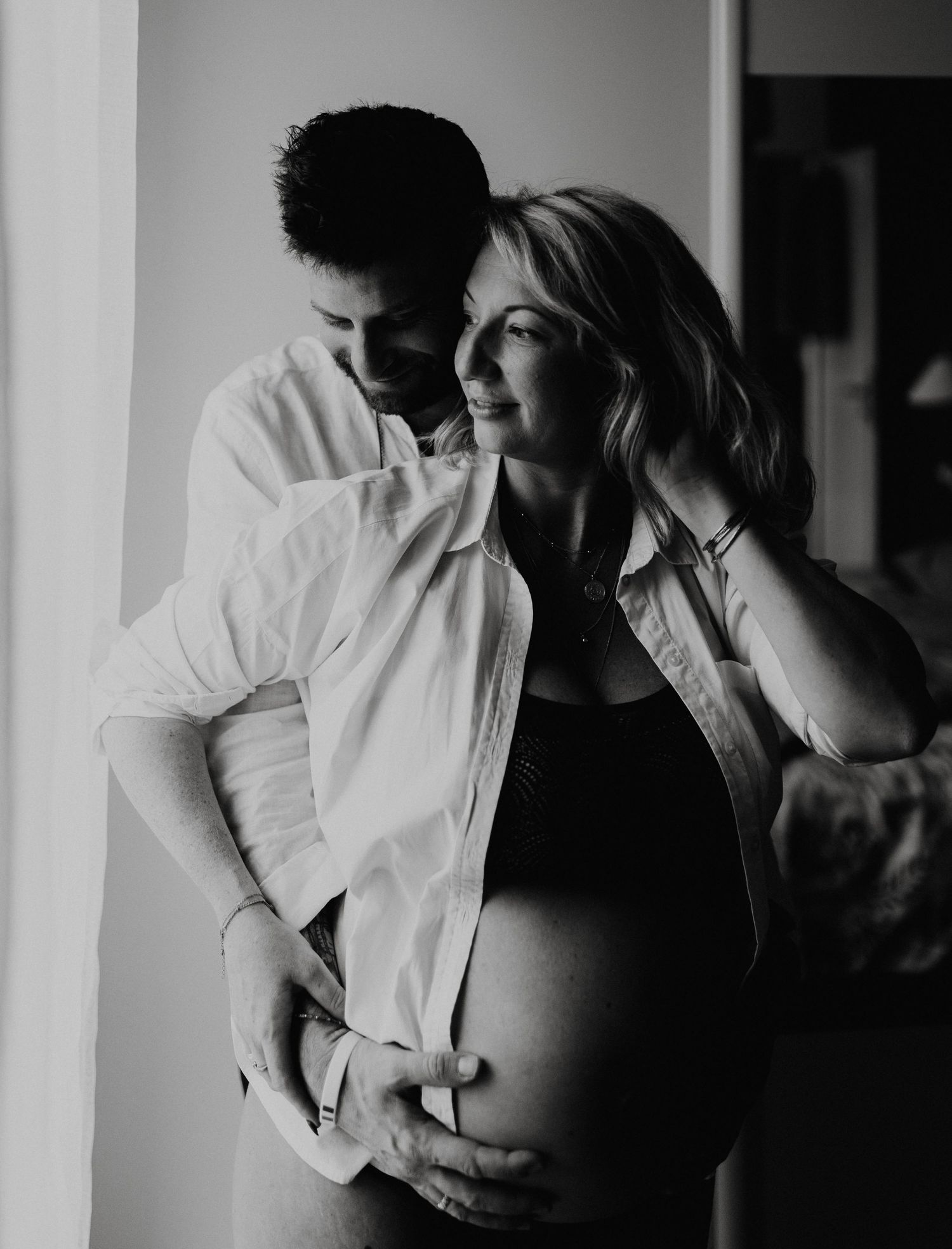 Intimate black and white maternity photo showing couple embracing near window.