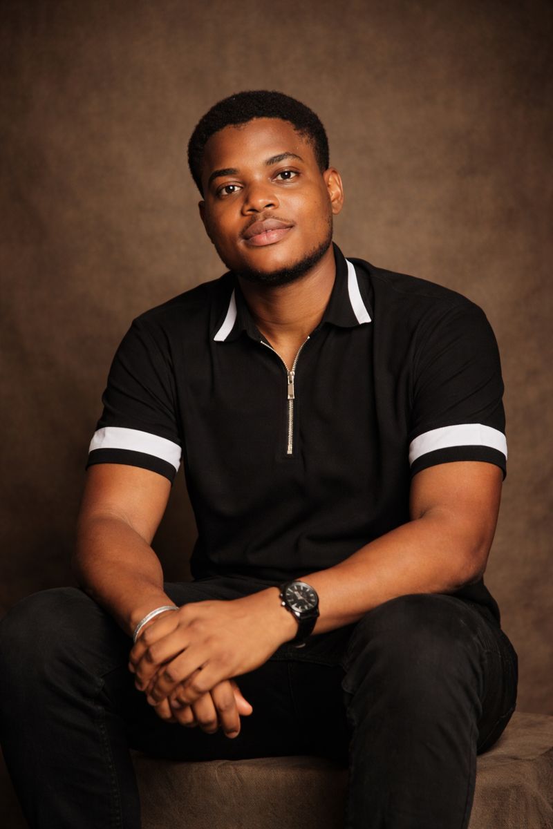 Professional studio portrait against brown backdrop showing person in black polo shirt with white trim sitting casually.