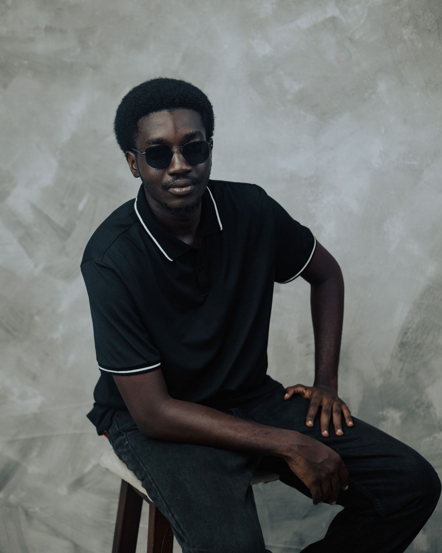 A person in a black t-shirt and sunglasses sits casually on a chair against a textured grey background.
