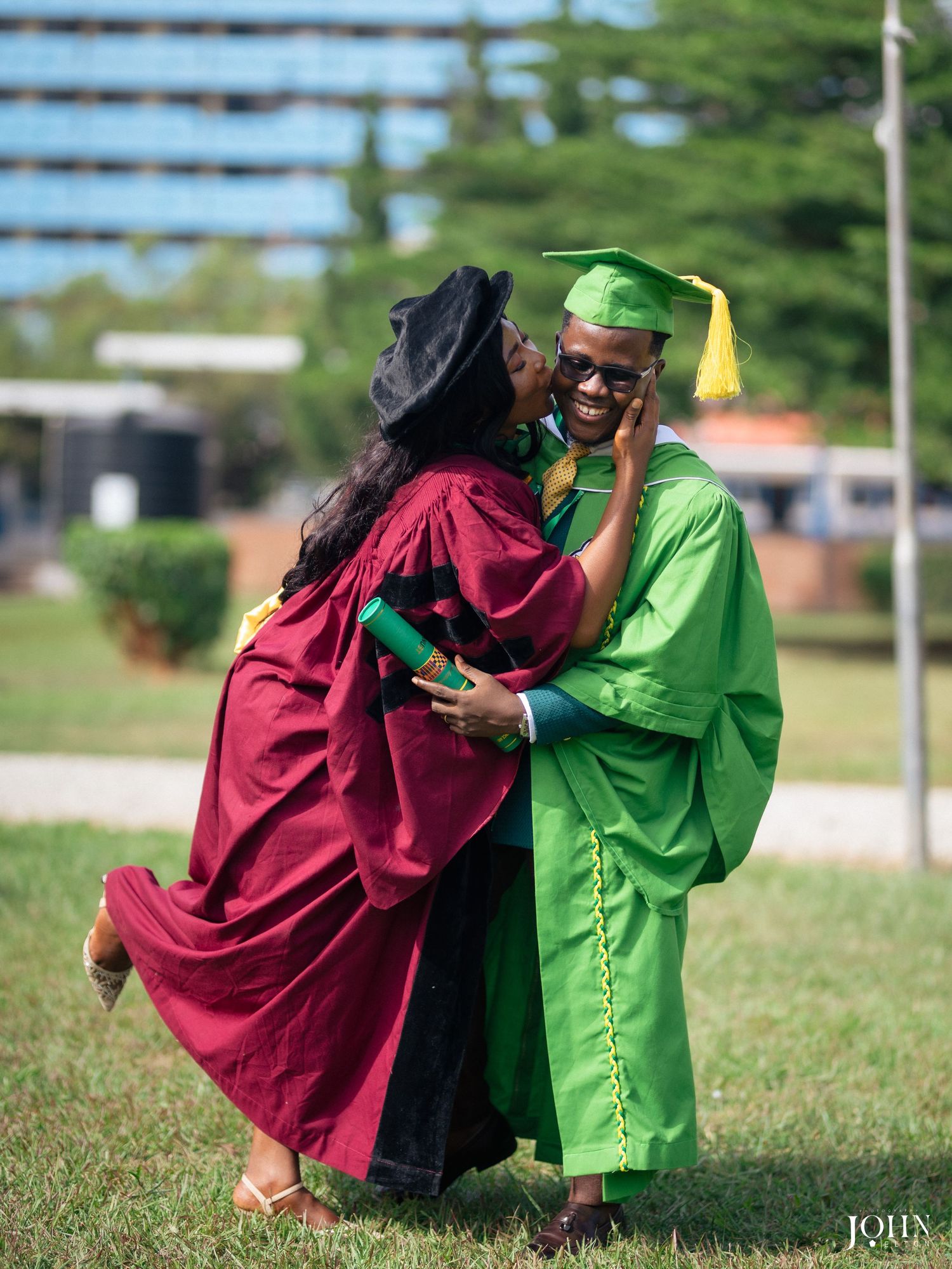 Two graduates in maroon and green regalia embrace during outdoor graduation celebration.