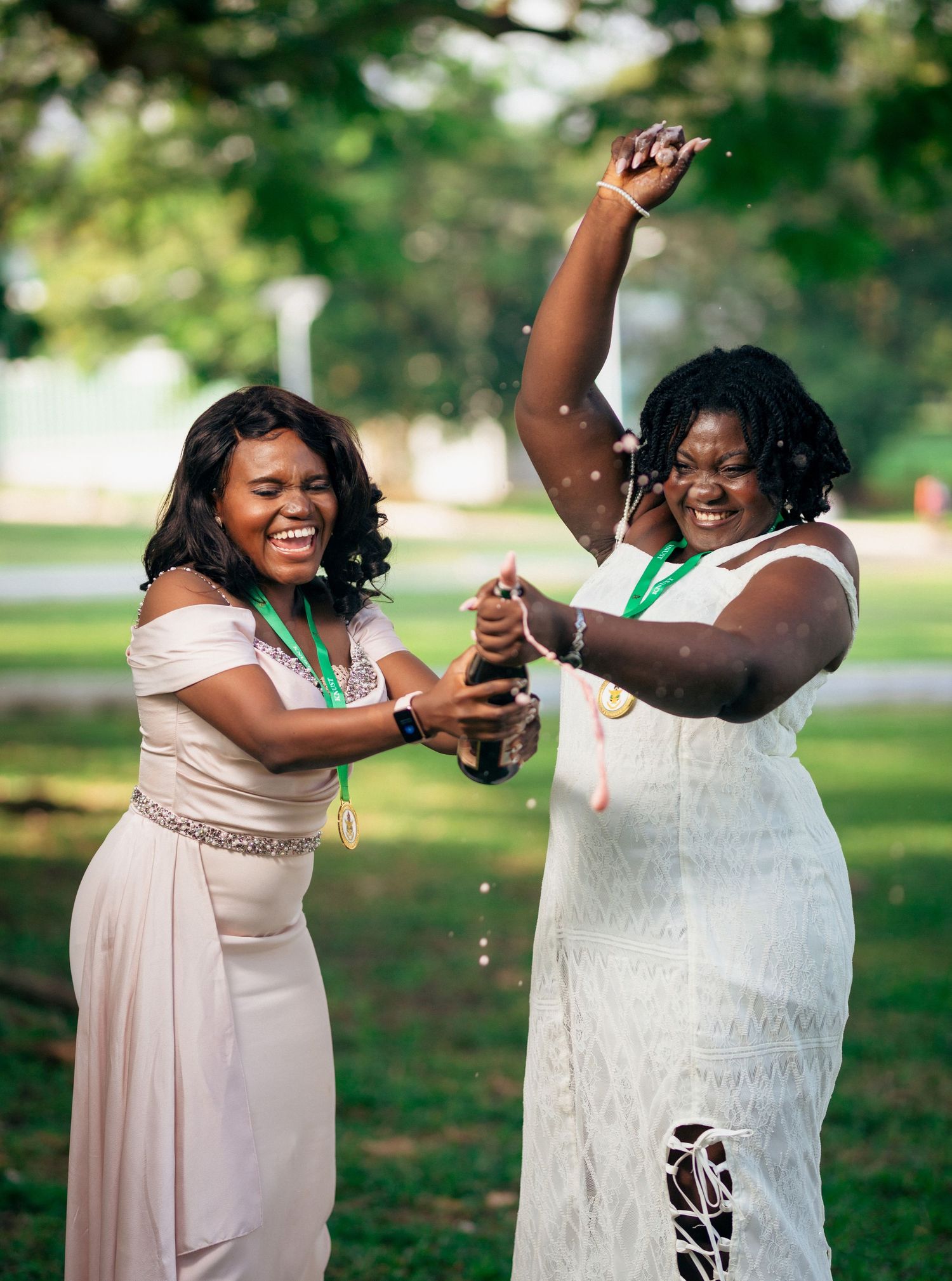 Graduates in light dresses pop champange, celebrate and pose together on a sunny day in a green park setting during their graduation.