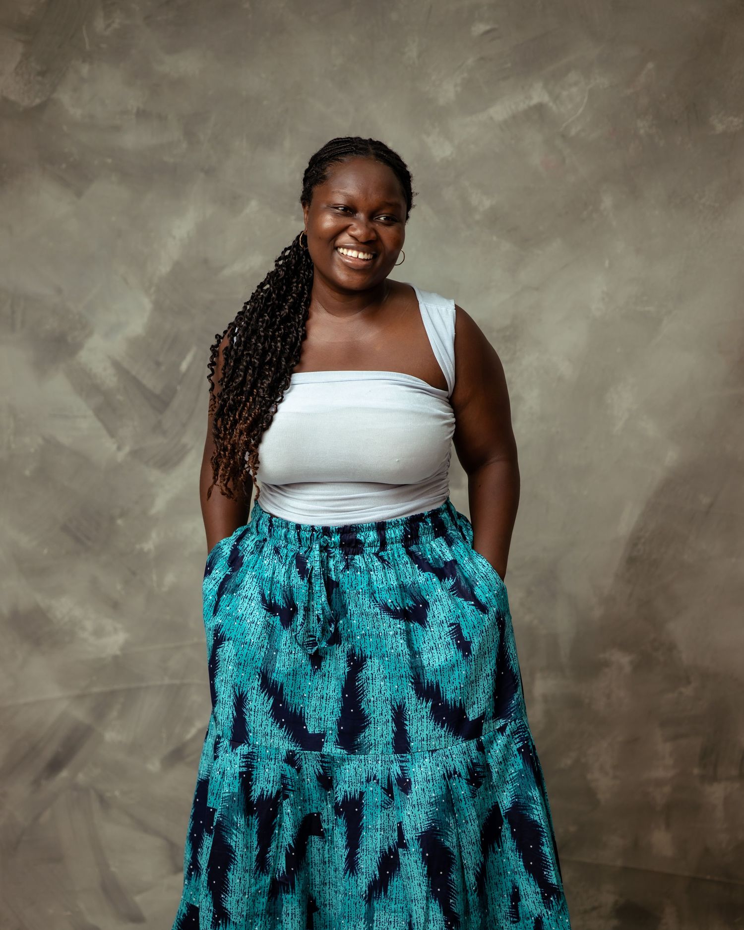 A person in a white tank top and turquoise patterned skirt poses against a textured gray background.