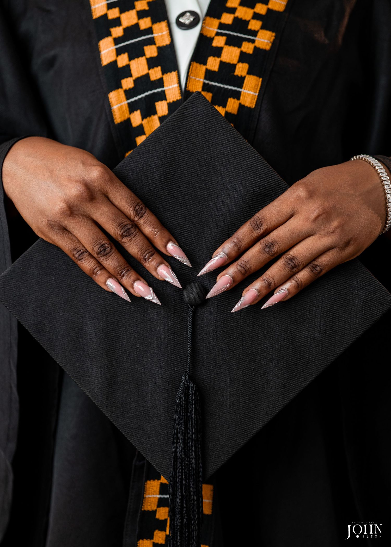 Close-up view of hands on black graduation gown with orange and black checkered stole and black tassel.