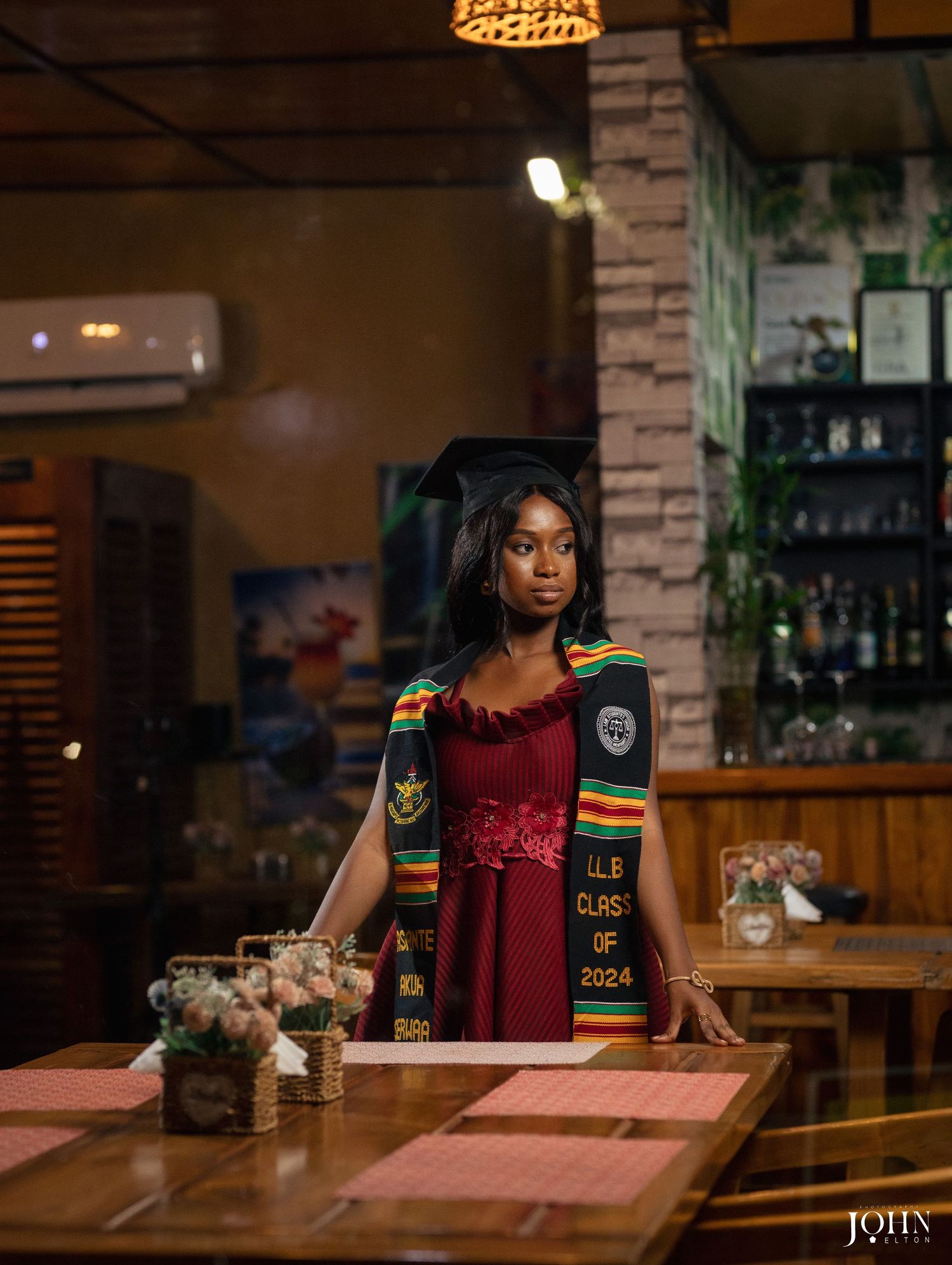 Graduate wearing red top and graduation stole poses in restaurant setting with ambient lighting.