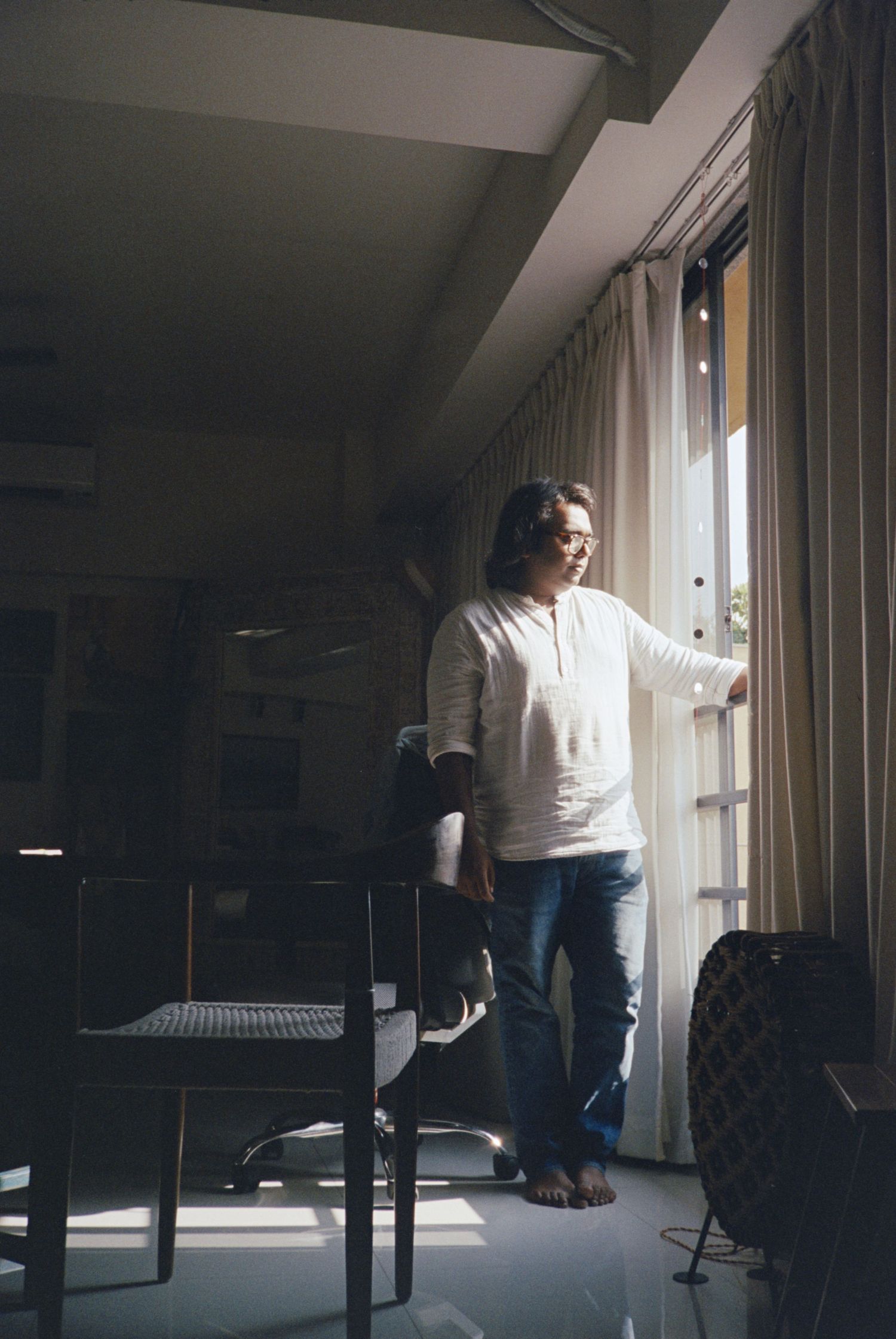 Person in white sweater gazing through window in moody interior scene with dramatic lighting and furniture silhouettes.