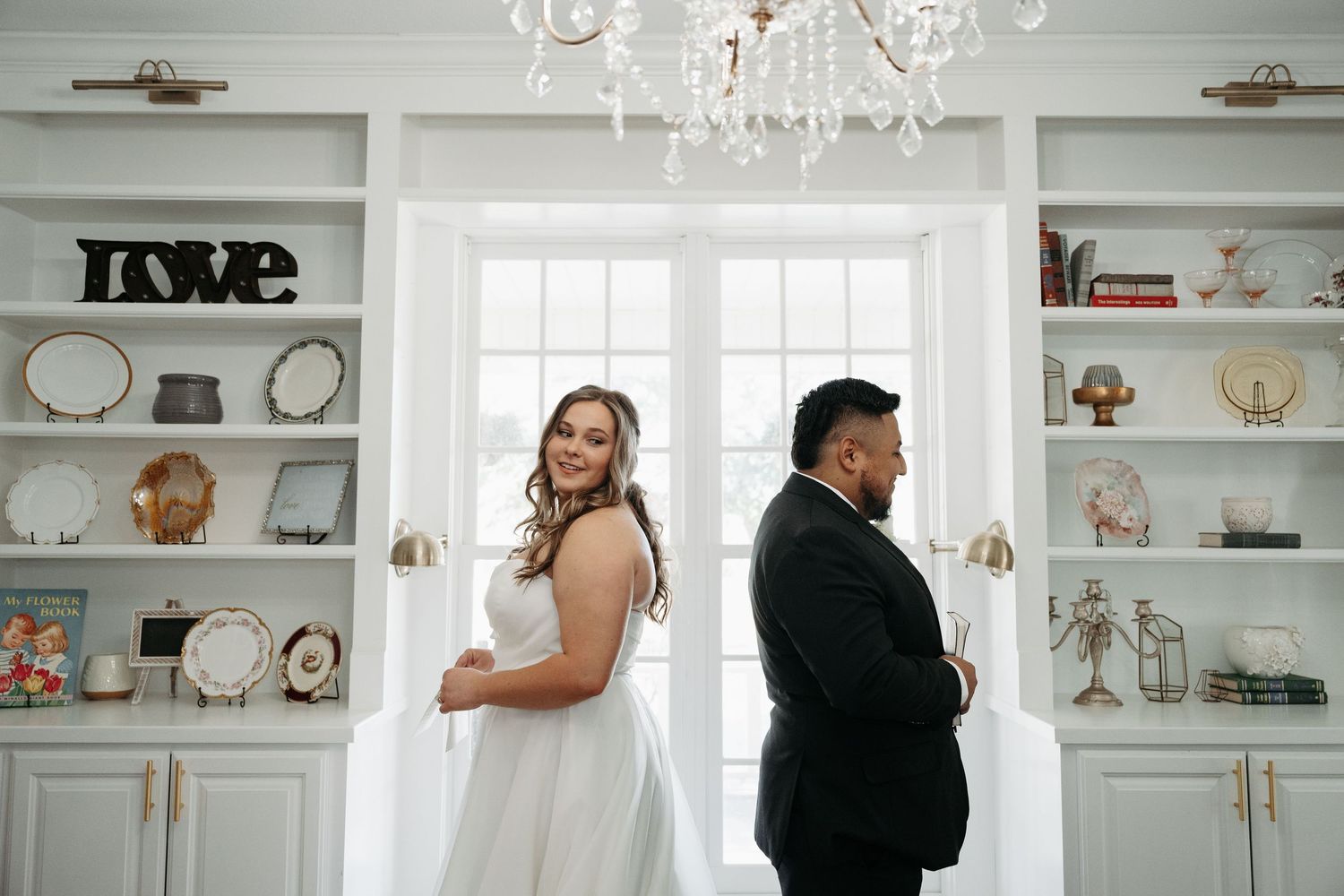 A romantic couple stands in an elegant white room with built-in shelves and a chandelier during a wedding photo session.