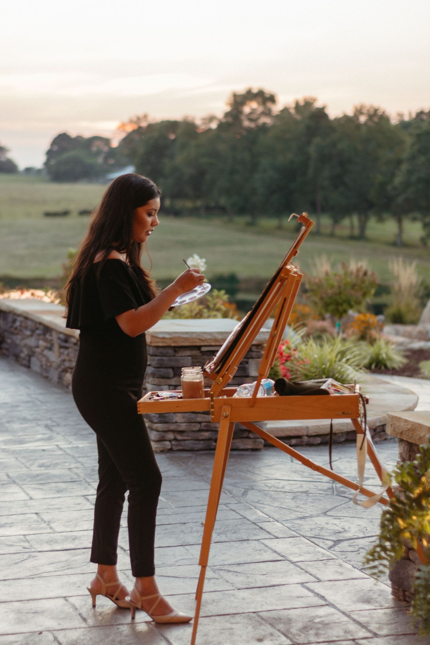 Artist working at a wooden easel outdoors on a stone patio during sunset.