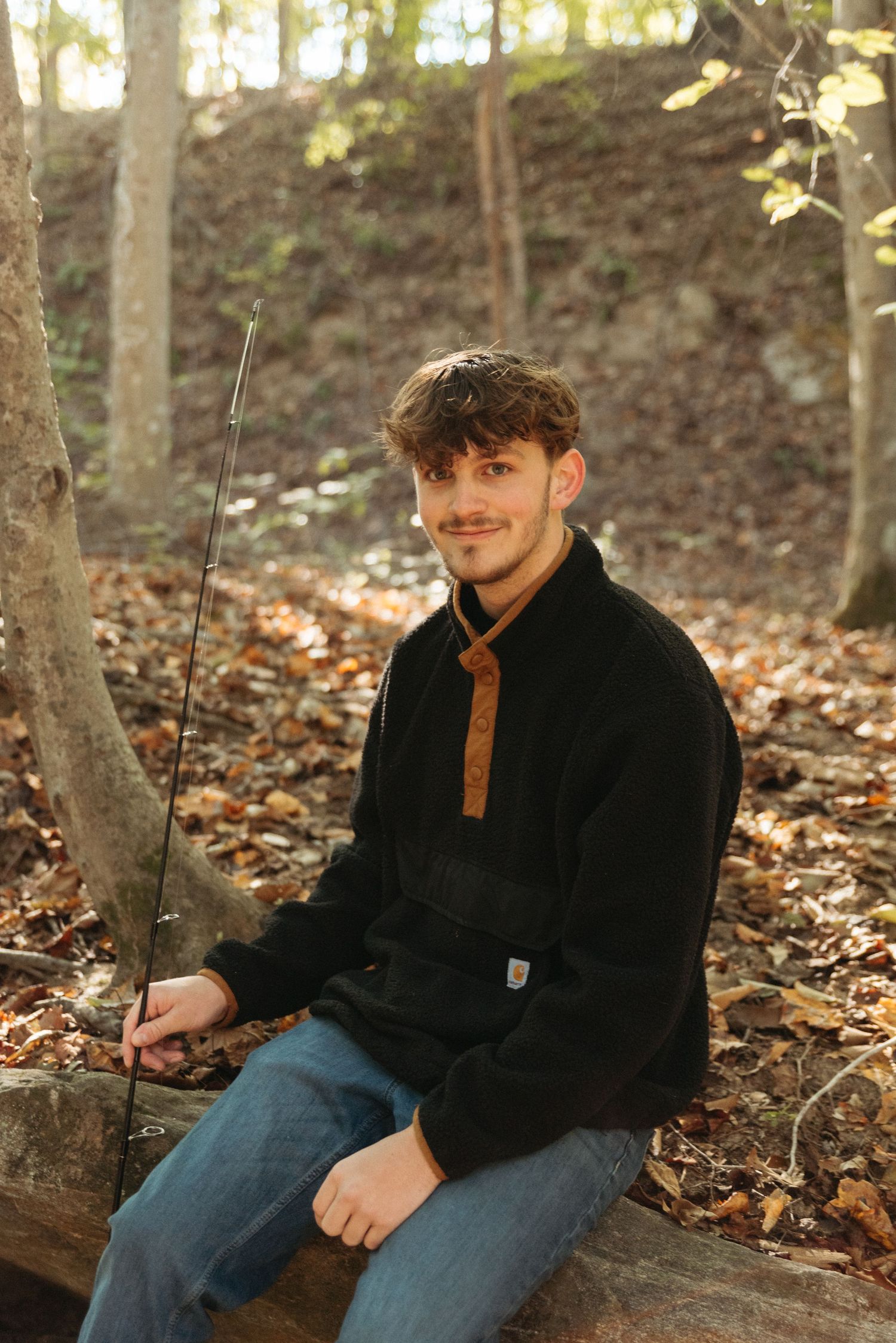 Person in black jacket sitting among fallen autumn leaves in a wooded area.