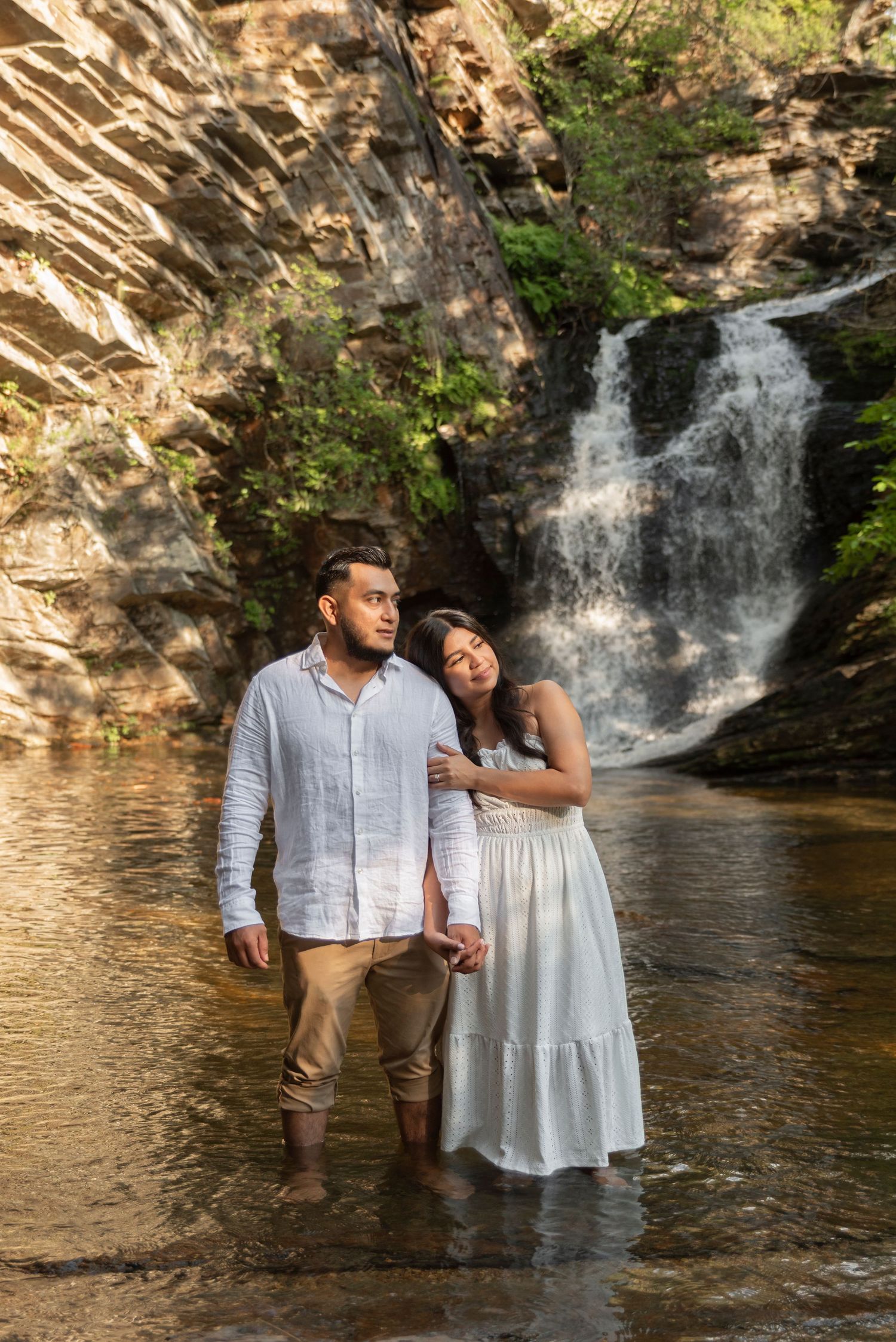 A couple stands together in front of a cascading waterfall in a scenic outdoor setting.
