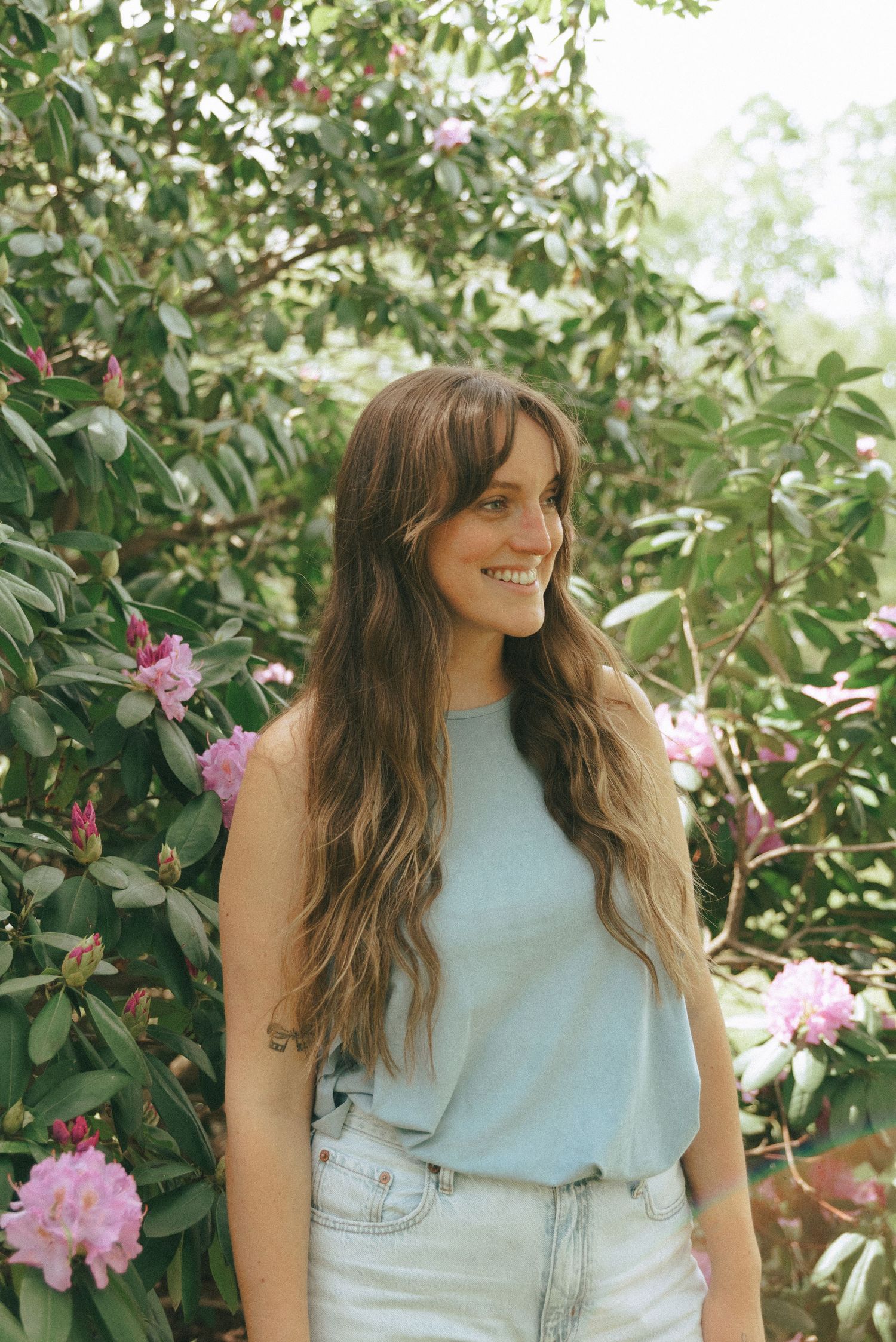 Woman in light blue outfit poses among blooming pink rhododendron flowers in a garden setting.