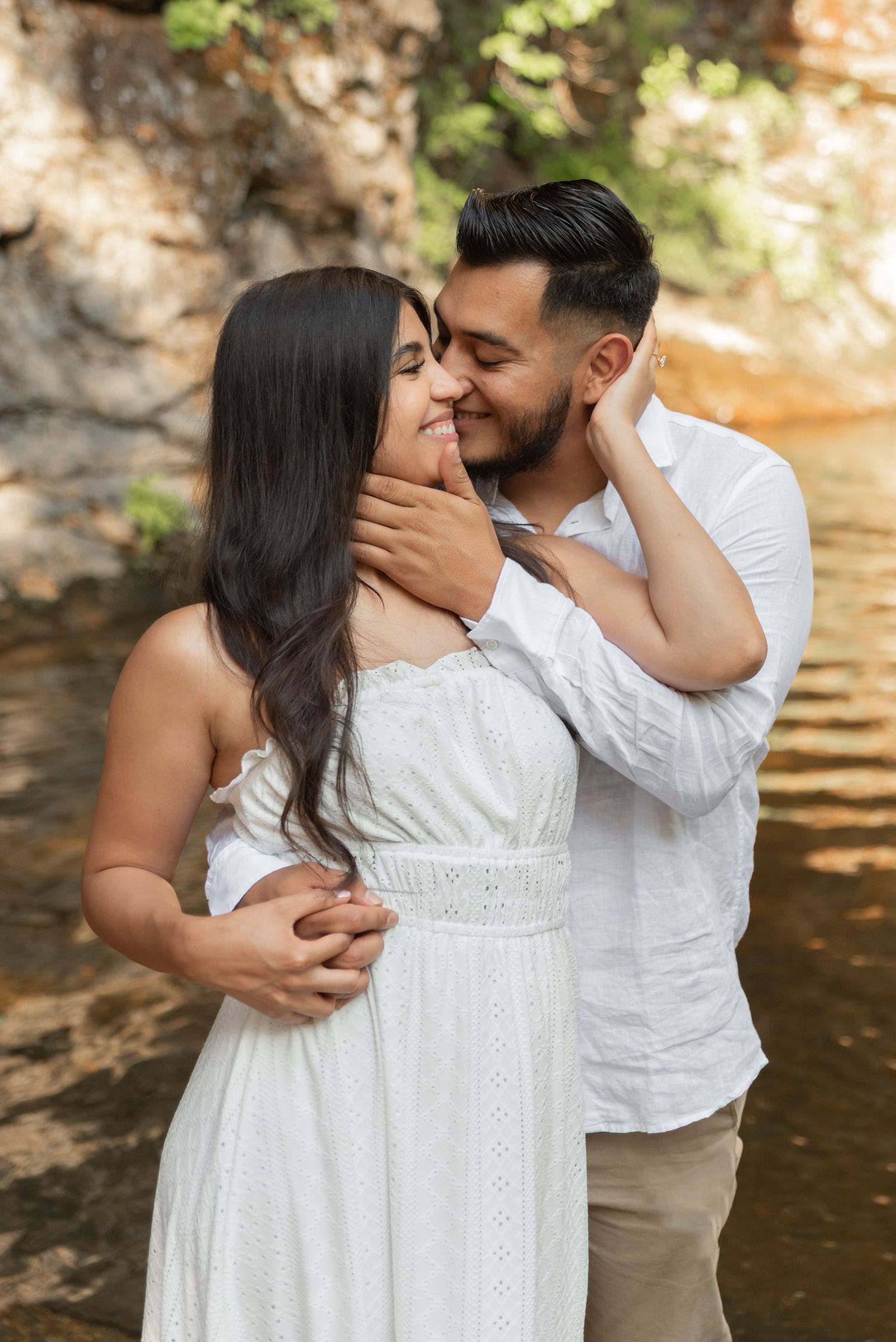 A romantic couple in white outfits share tender kisses by a serene waterfront in a series of intimate photos.