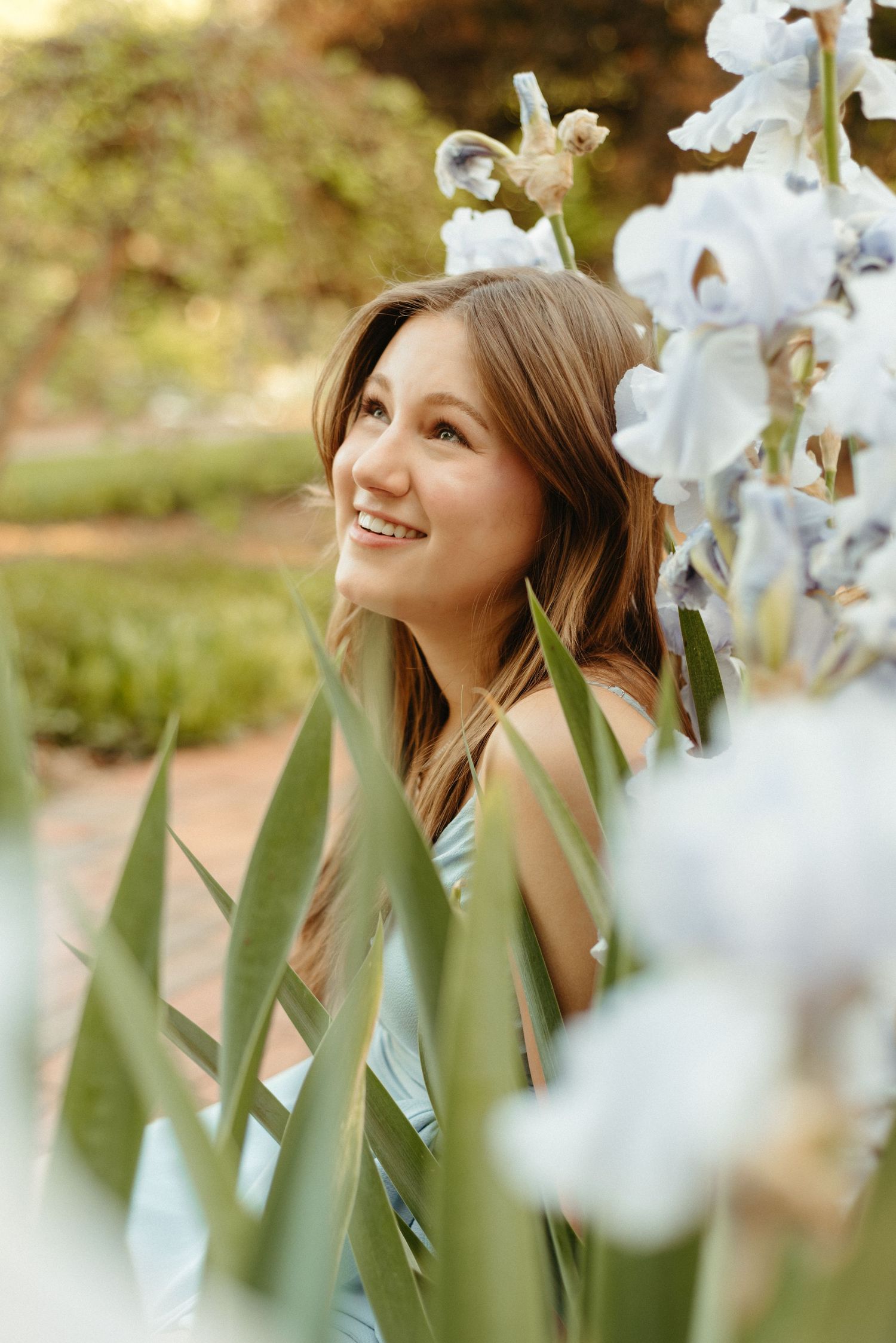 A person smiles joyfully while posing among blooming white iris flowers in a sunlit garden setting.