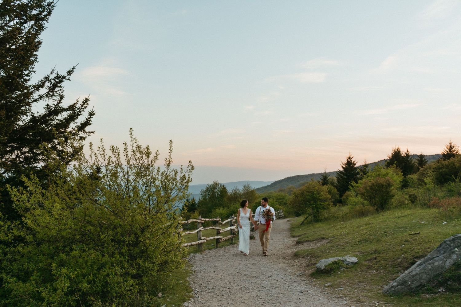 Scenic mountain trail view with distant figures walking along rustic fence at sunset.
