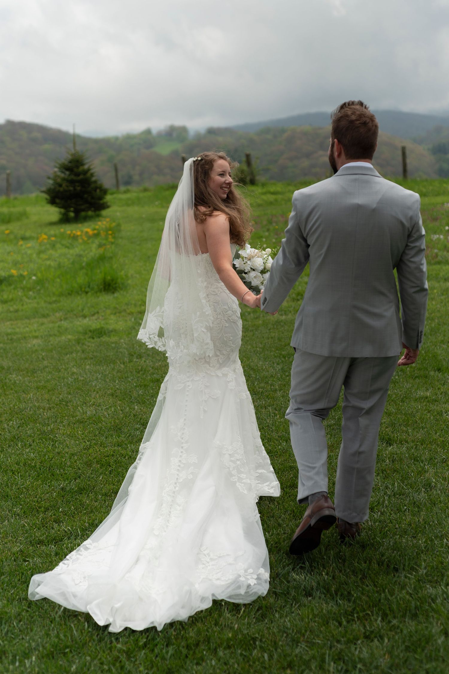Wedding couple walking hand in hand across rolling green hills with mountains in background.