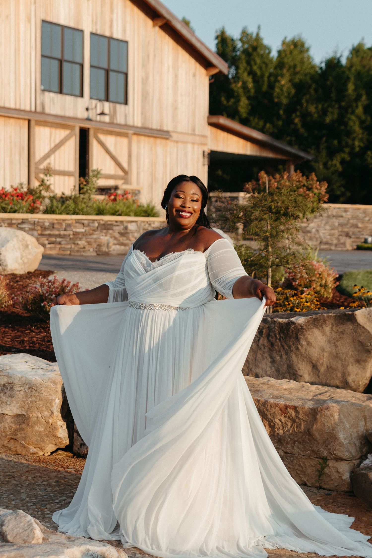 A bride in a flowing white dress poses joyfully against a rustic outdoor backdrop.