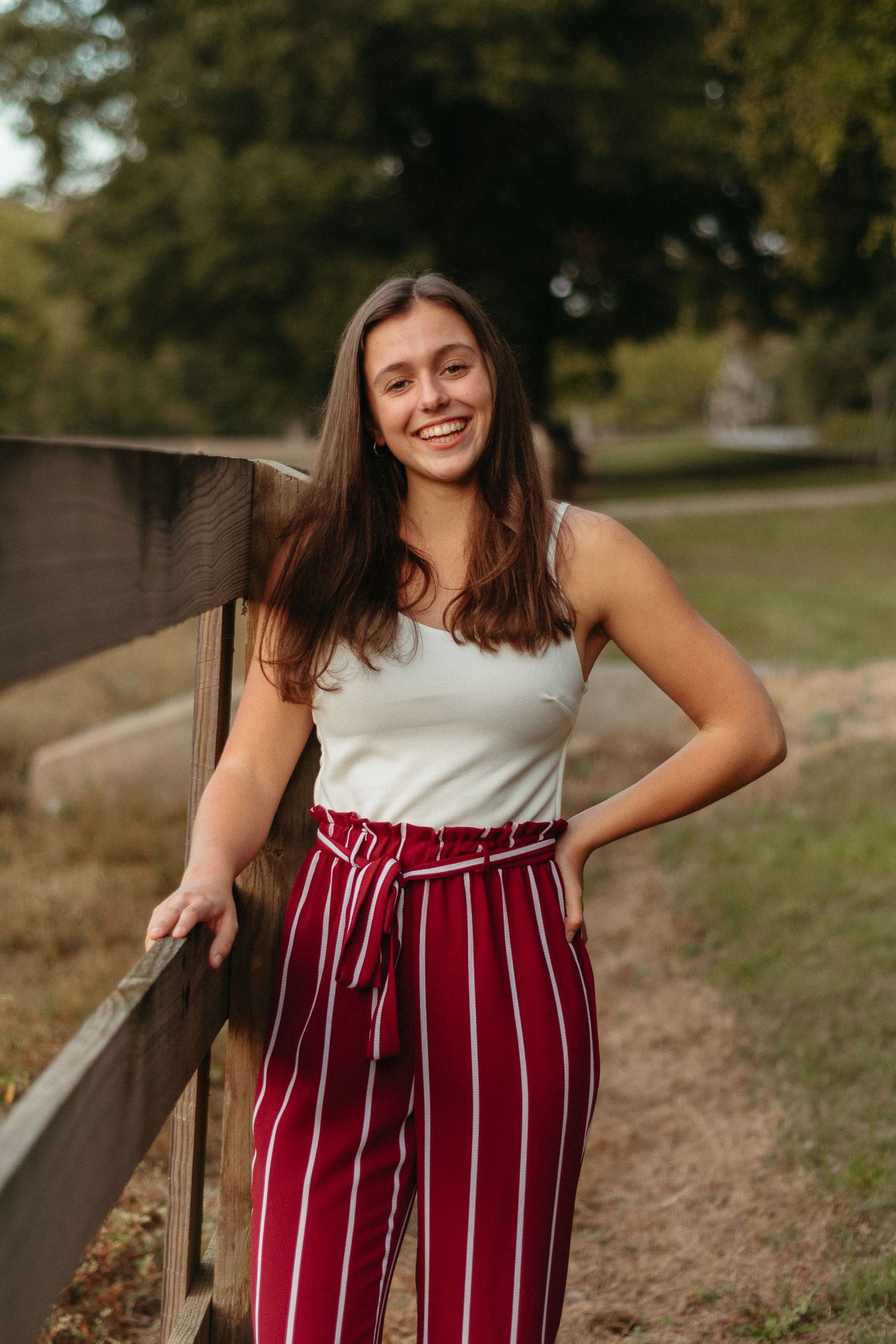 A person in a white top and burgundy striped pants poses outdoors against a wooden fence in natural lighting.