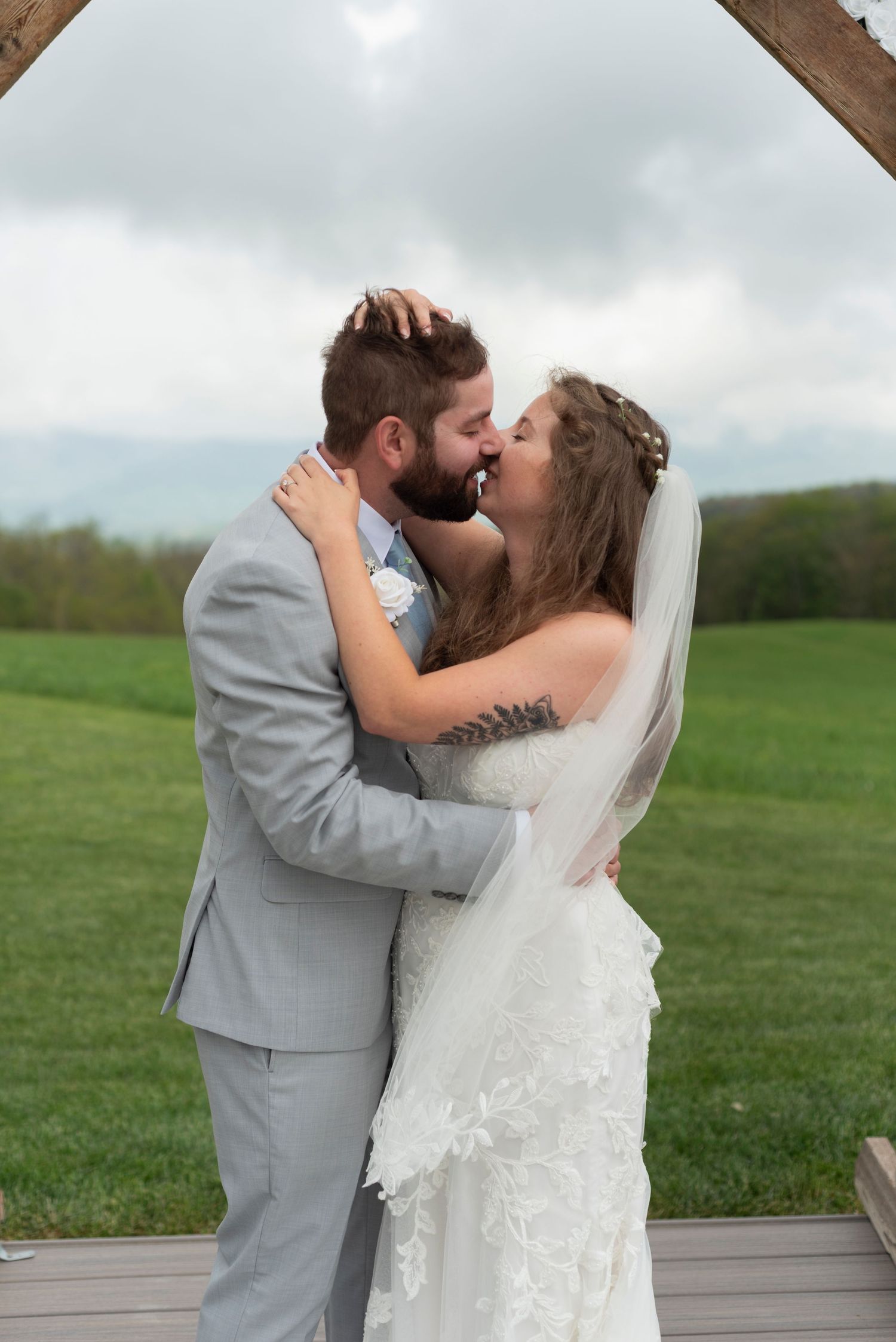 Couple in wedding attire sharing their first kiss on an outdoor deck with green hills in background.