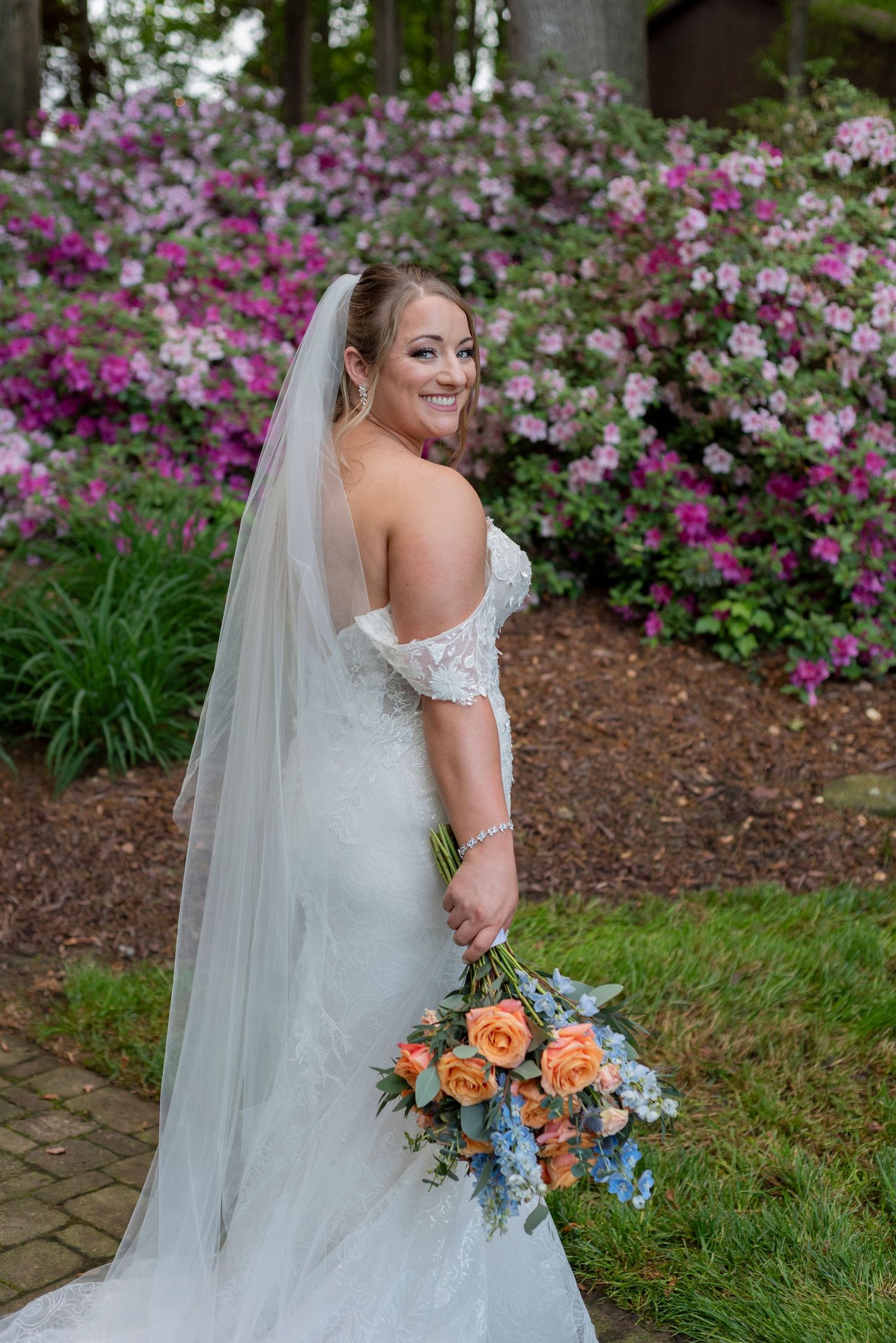 Bride in lace gown poses with colorful bouquet against backdrop of pink and white flowers.
