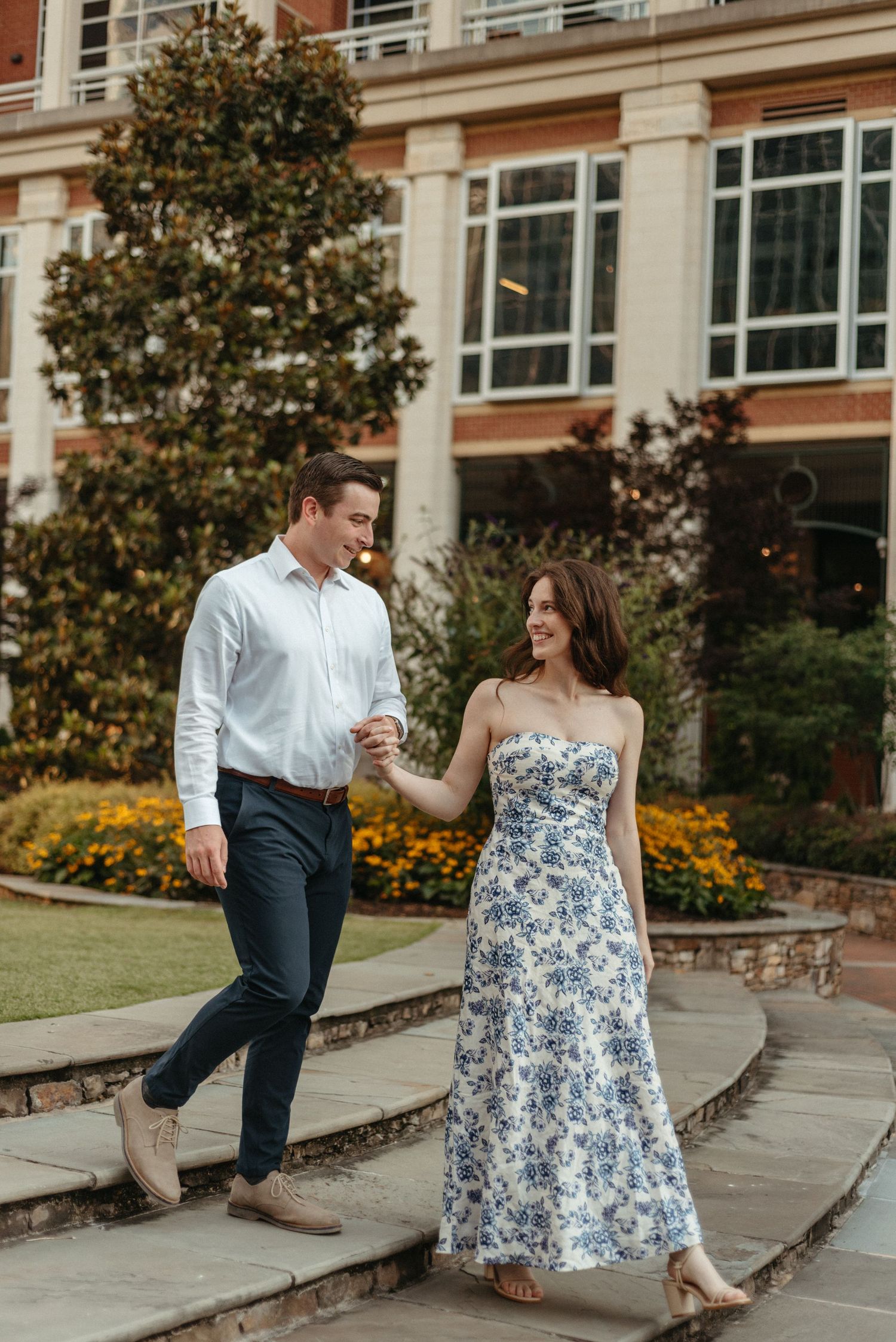 A couple sits and laughs together on stone steps outside a brick building with autumn foliage in the background.