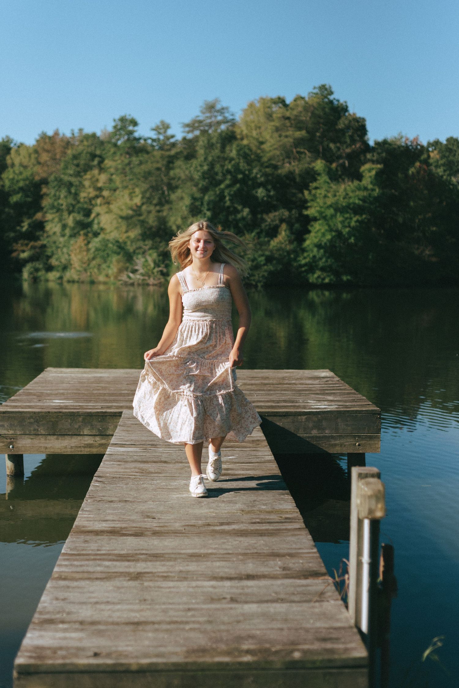A person in a white dress walks along a wooden dock over a serene lake.
