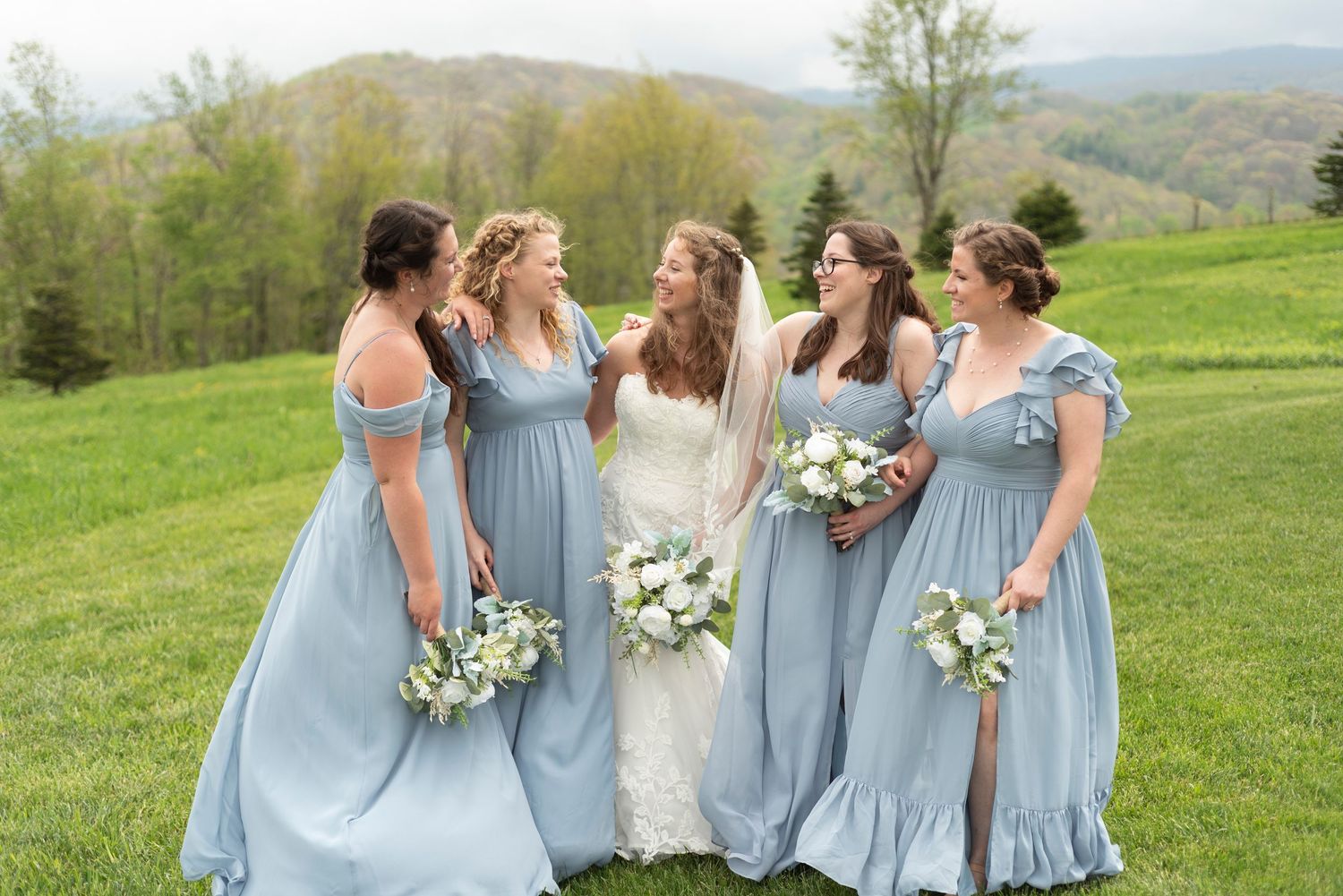 Bridesmaids in light blue dresses hold bouquets while standing in a row on a grassy hillside.