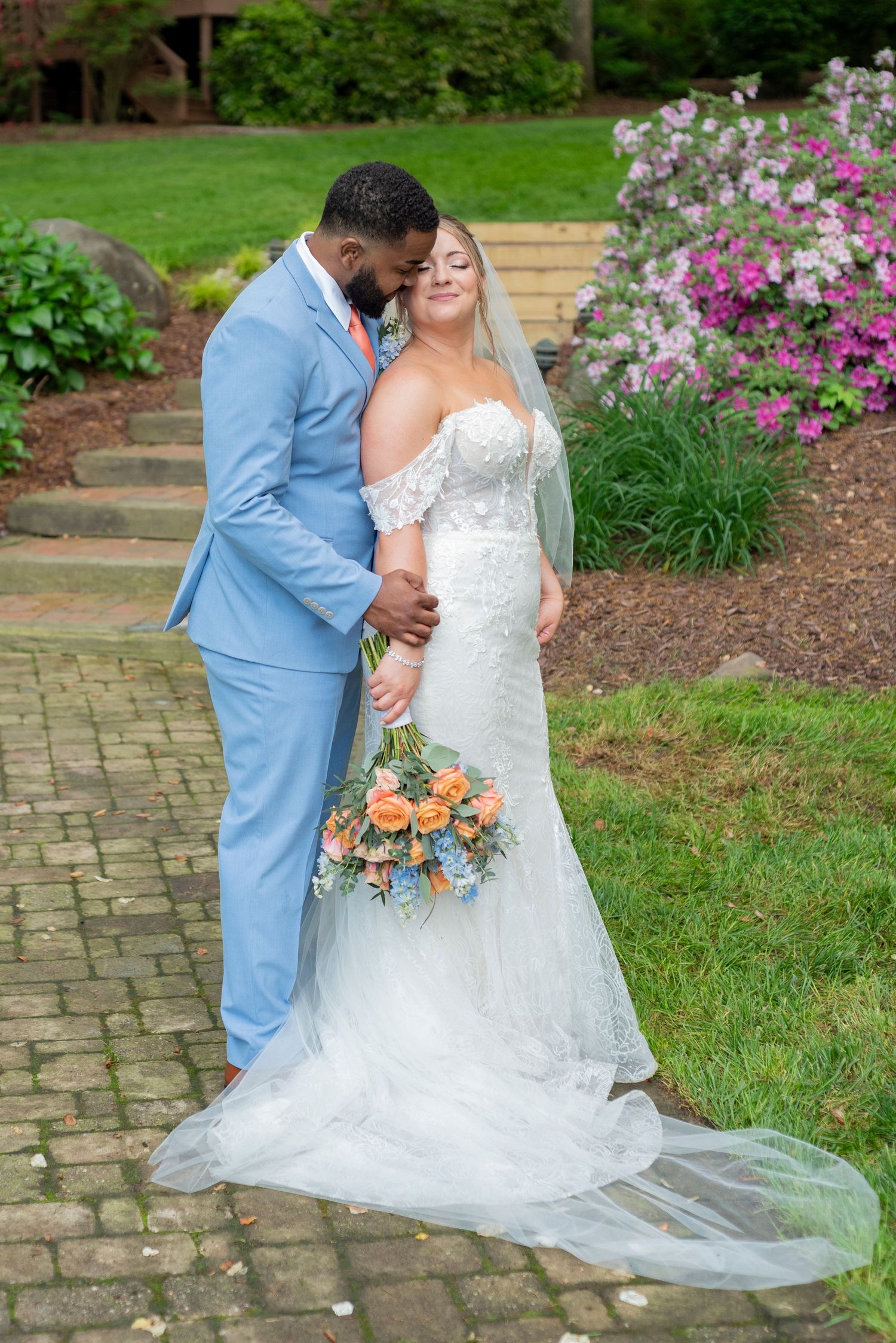 Couple in wedding attire shares intimate moment on stone pathway surrounded by flowering gardens.