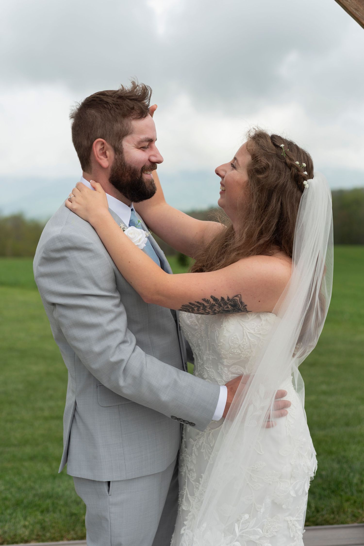 Newlyweds embrace in grey suit and white dress against cloudy sky backdrop.