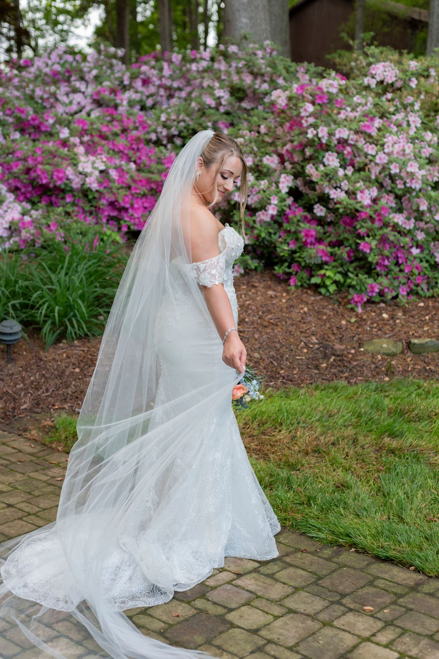 Bride in white gown holds vibrant bouquet while posing by garden of pink and white flowers.