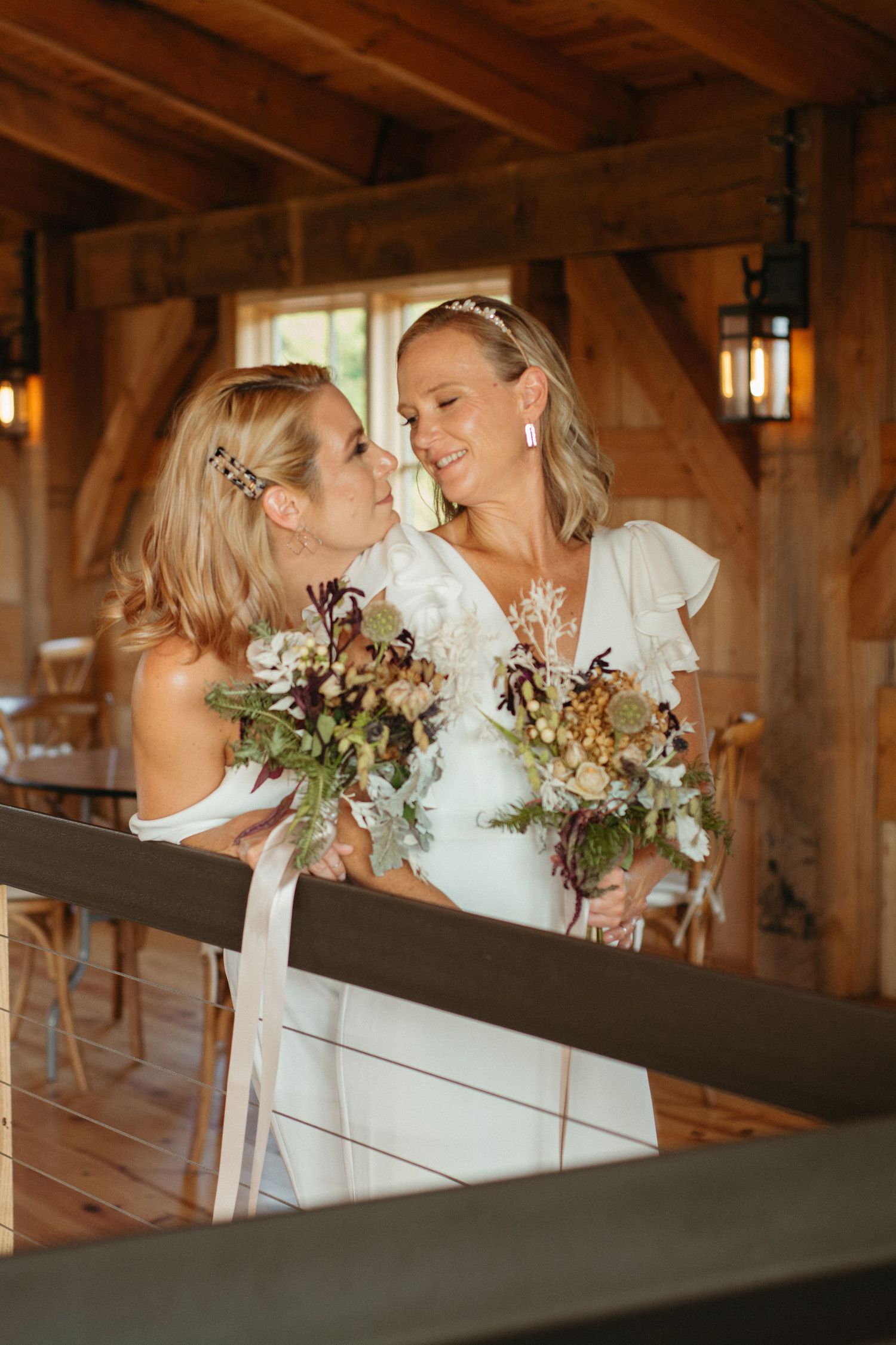 Rustic barn interior with wooden beams where two people in white dresses share an intimate moment.