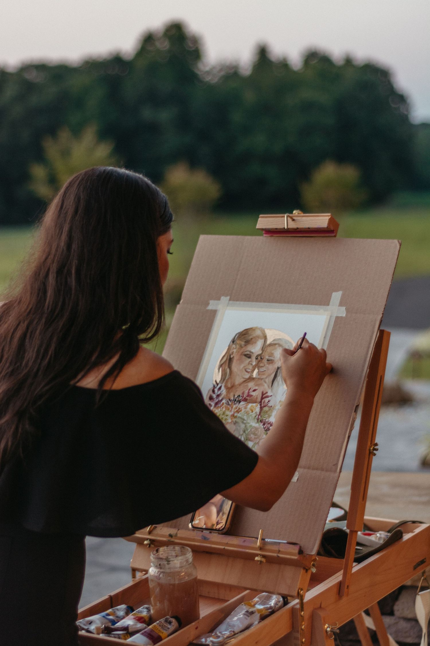 Artist painting at an outdoor easel with artwork visible on canvas during golden hour.