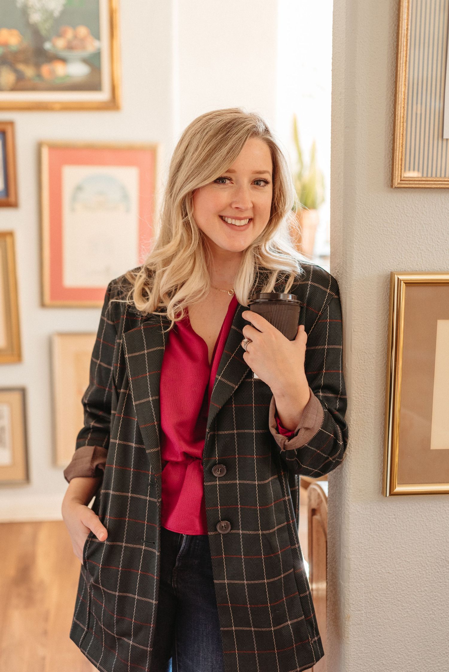 Person in windowlit gallery wearing plaid blazer and red top poses between framed artwork.