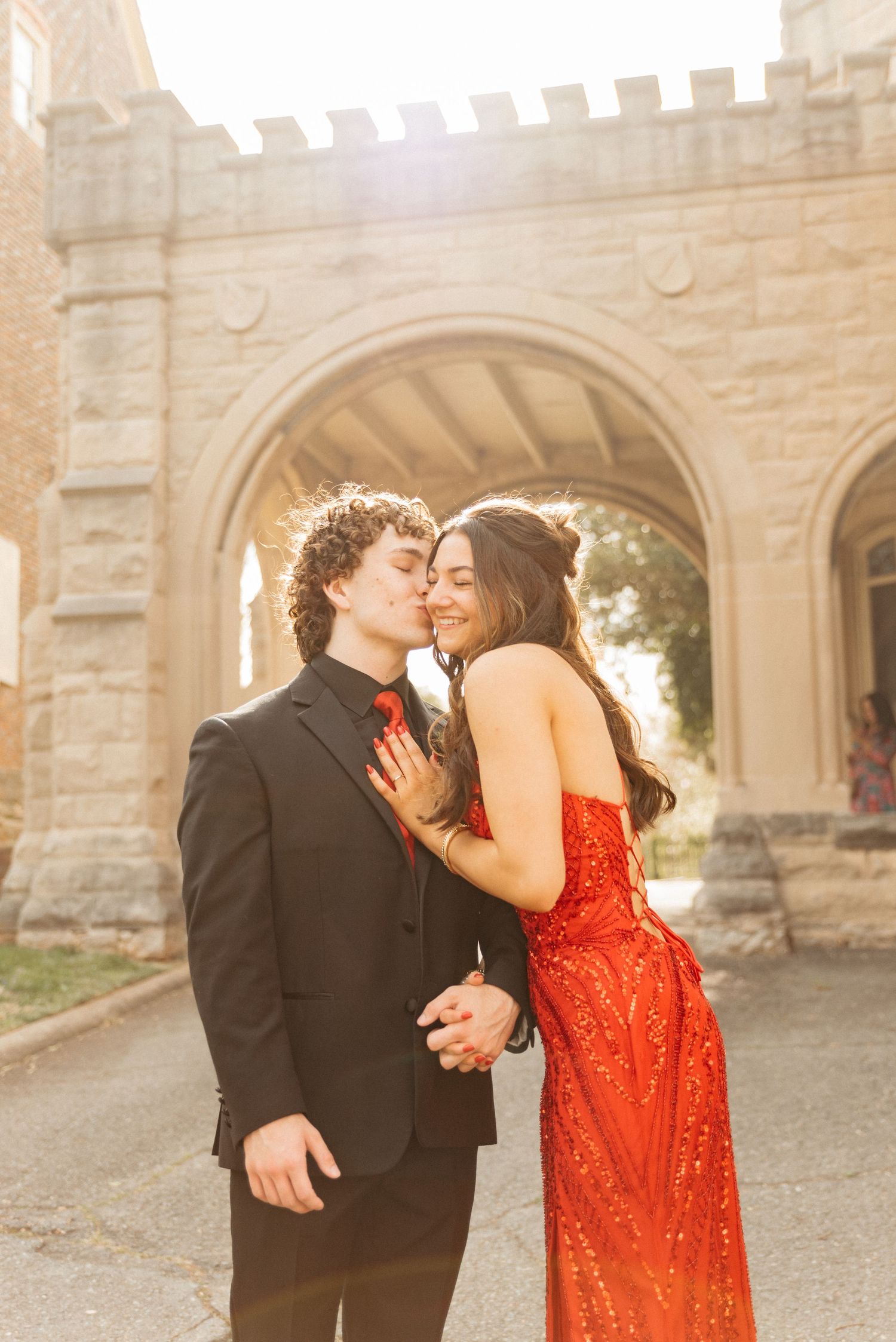 A couple in formal attire share an intimate moment under a stone archway at sunset.