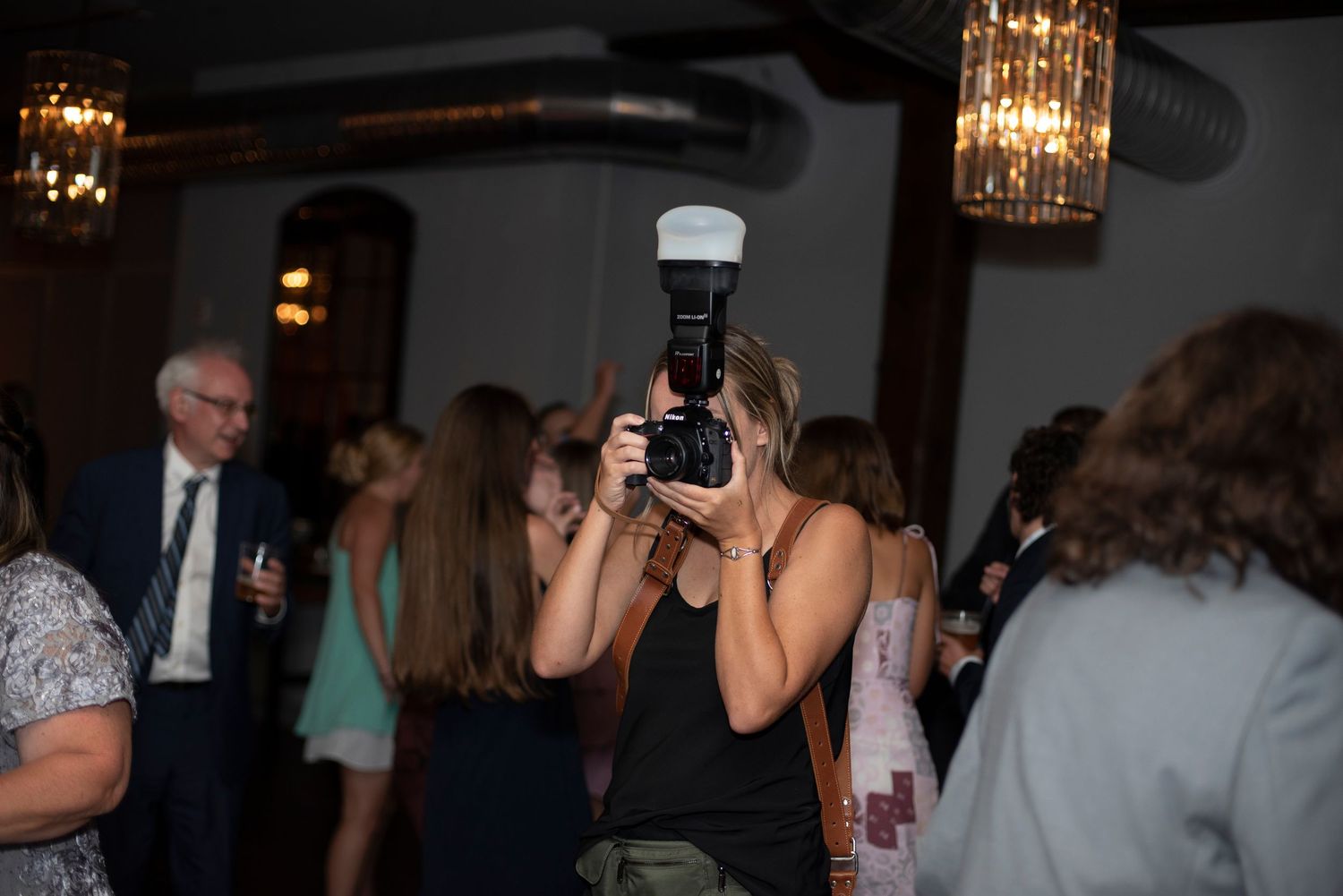 People dancing and taking photos at a dimly lit party or celebration venue with crystal chandeliers overhead.