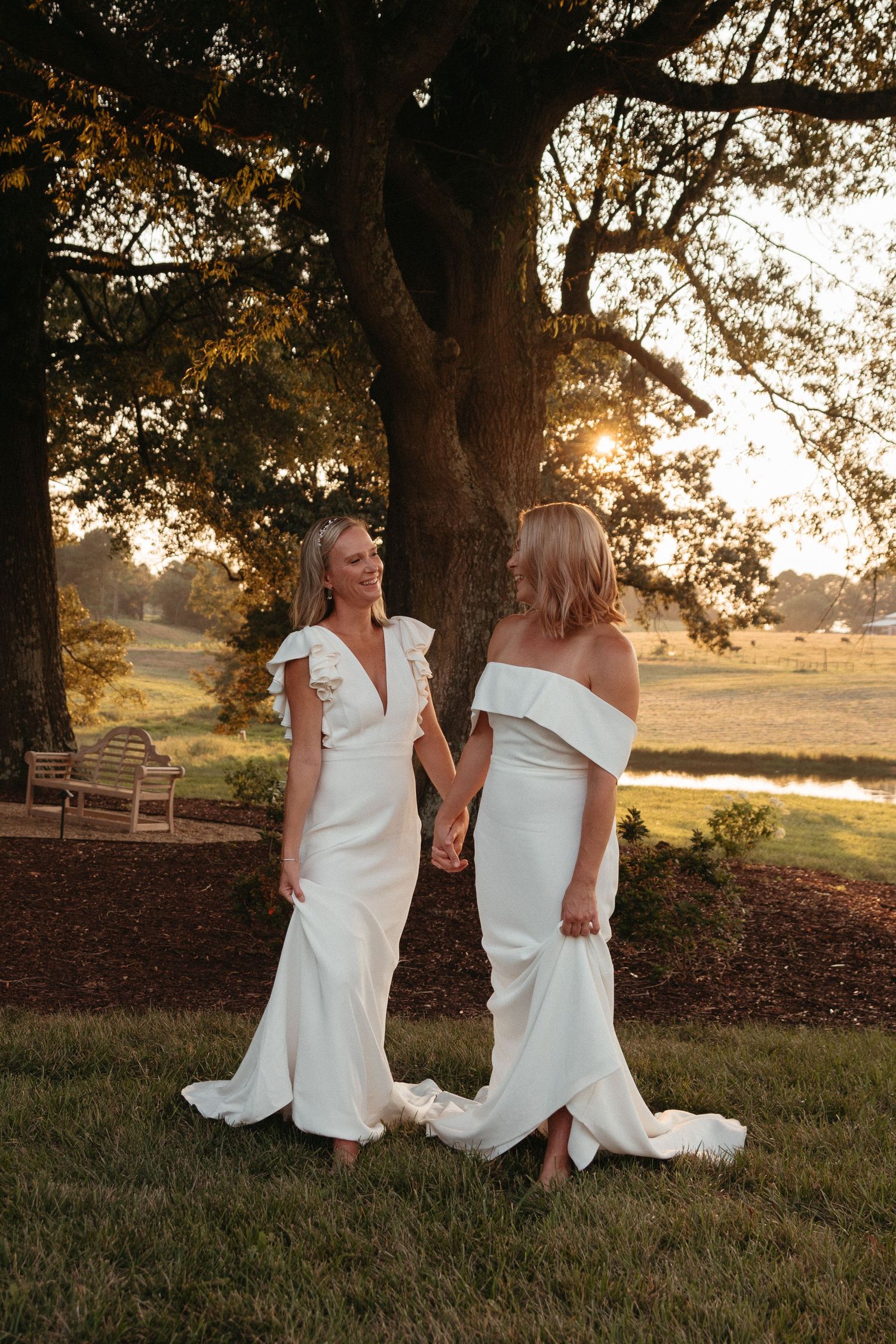 Two people in white dresses share a joyful moment on a grassy hill during golden hour sunset.