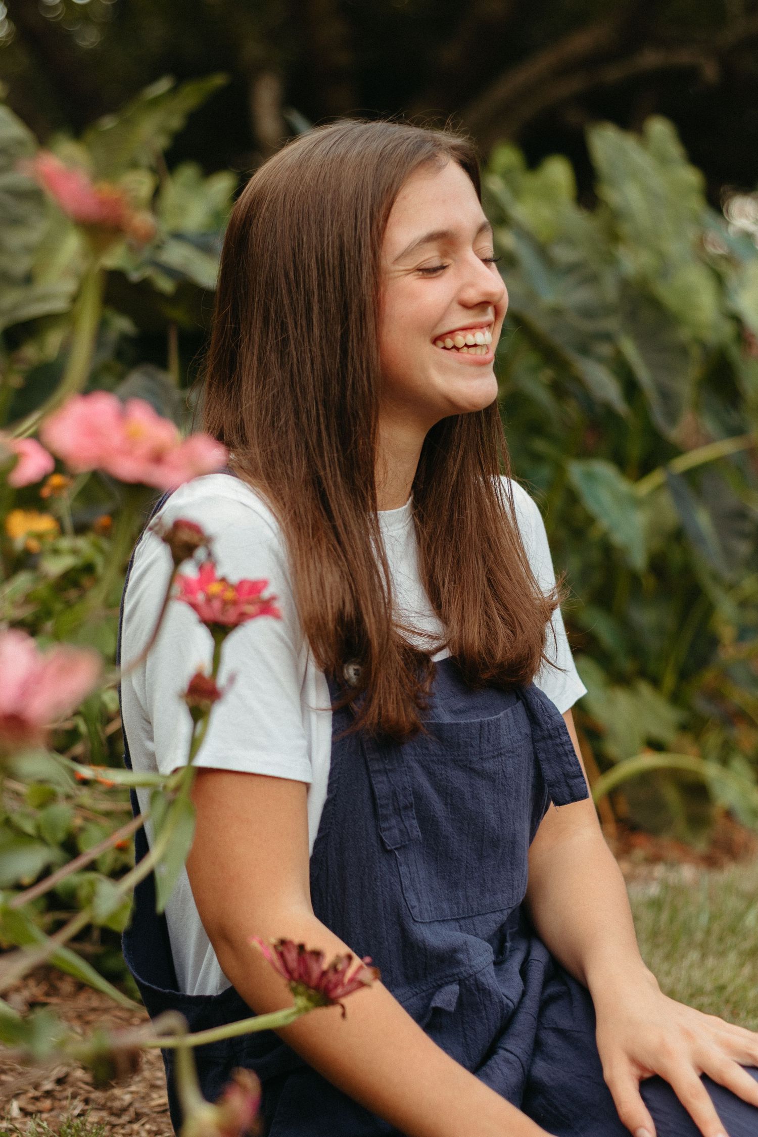 A person in a navy blue pinafore dress and white shirt sits outdoors surrounded by flowers and greenery.