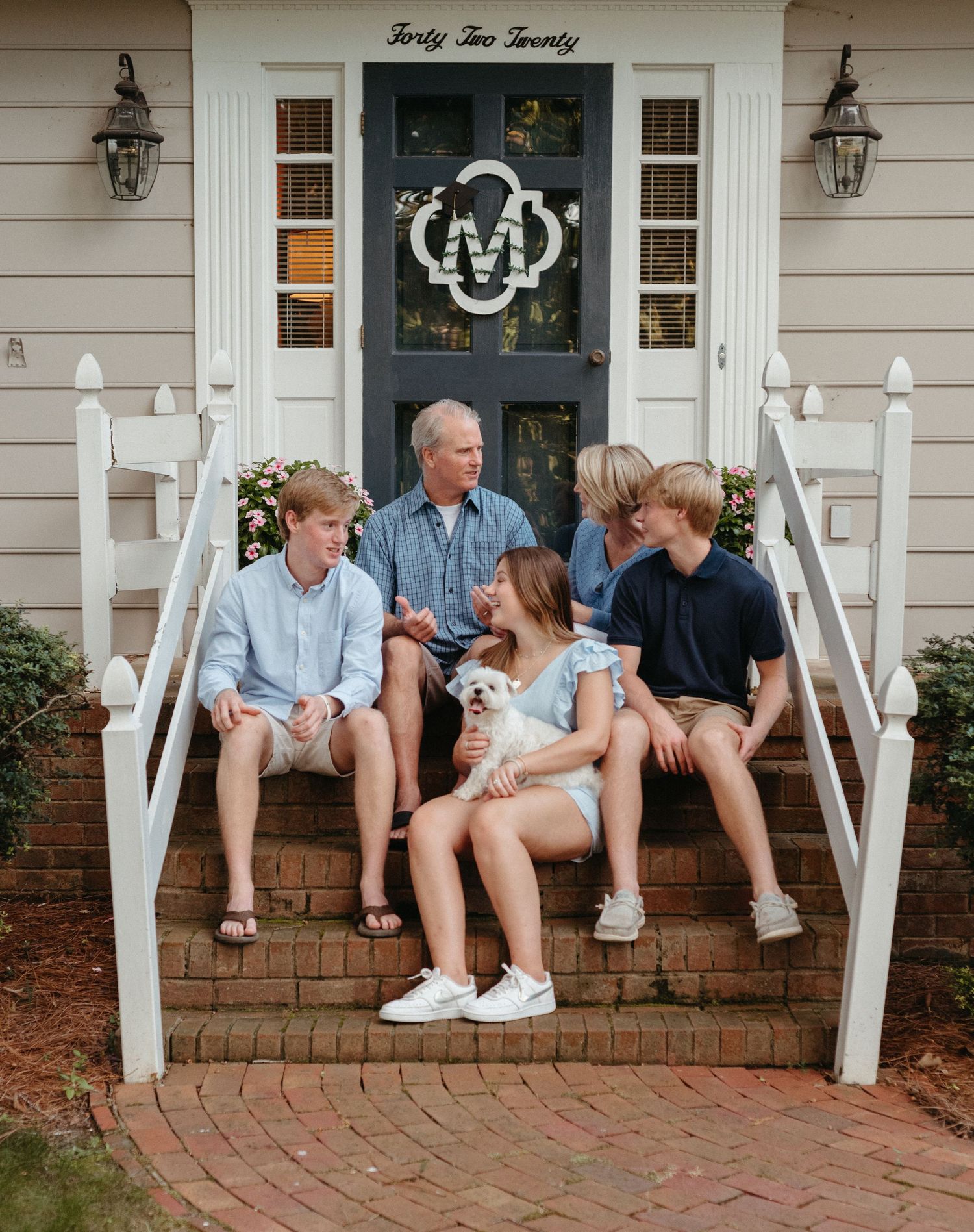 Family sits together on white porch steps of traditional home.